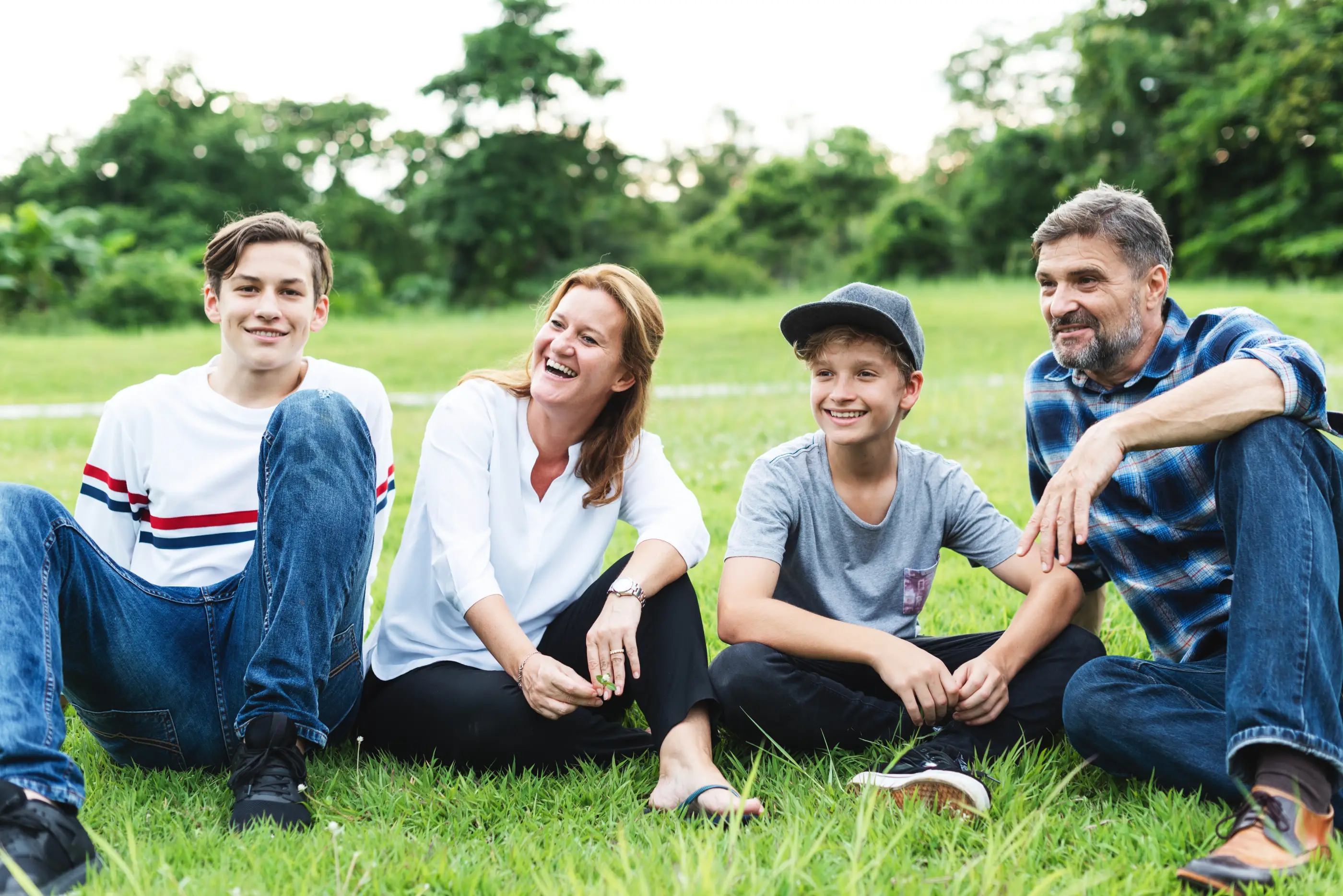 Smiling family of four sitting on grass outdoors with trees in the background.