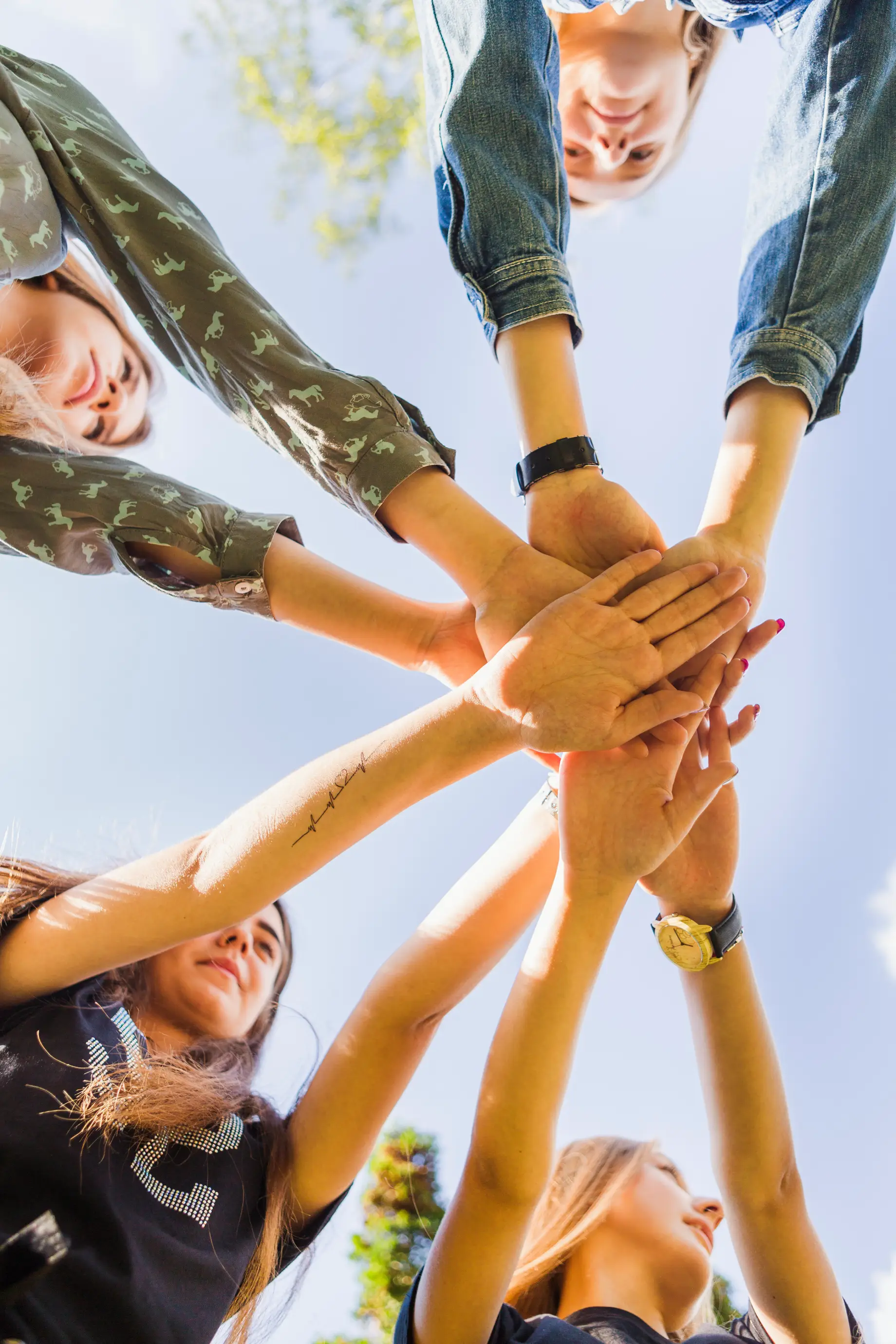 Four friends standing in a circle stacking their hands together outdoors under a clear blue sky.