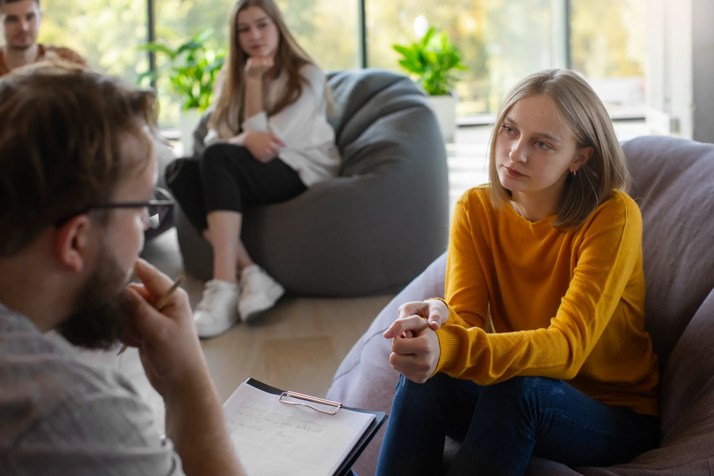 Young woman in mustard sweater attentively listens to a man holding clipboard during group therapy.