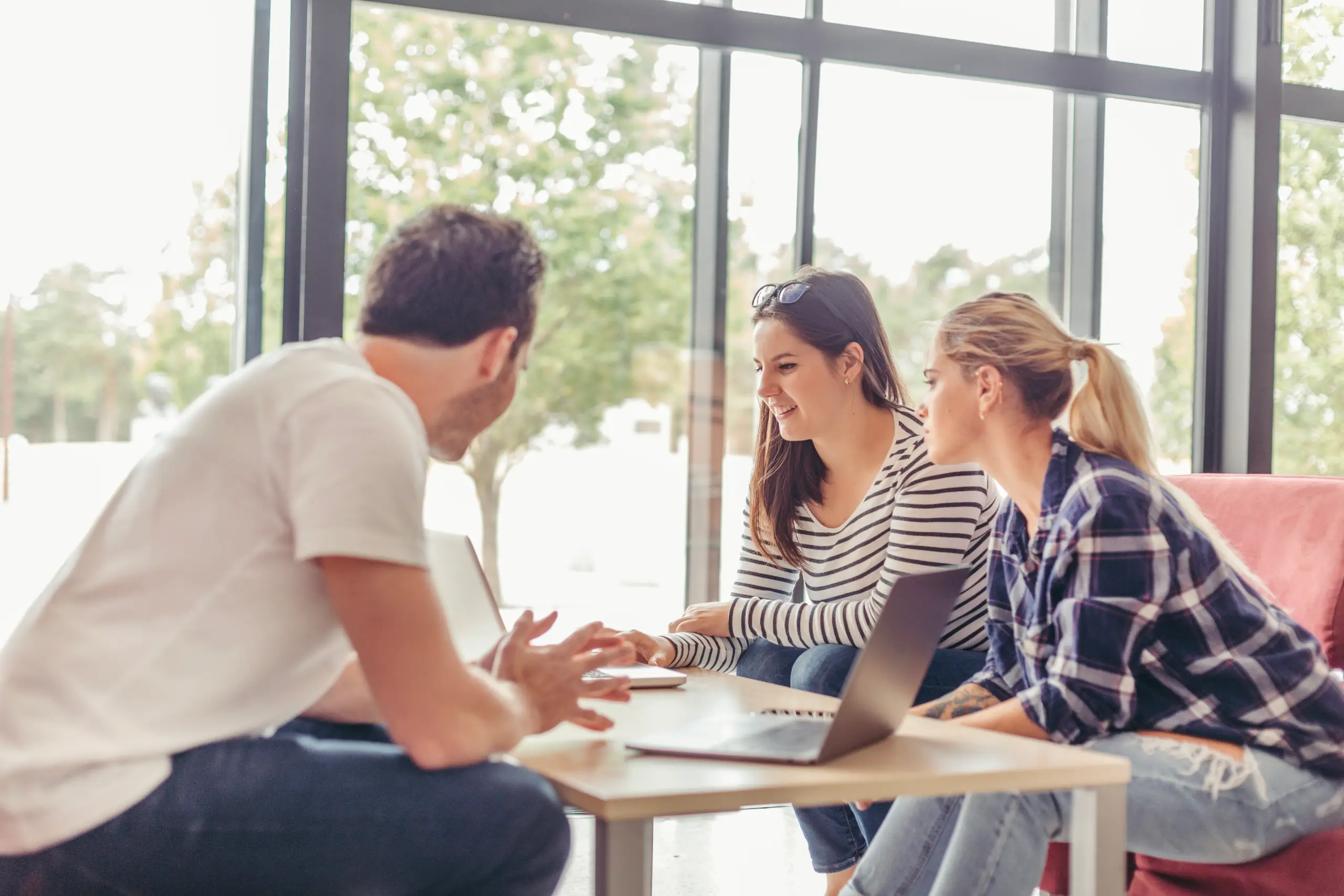 Three people sitting by a table with laptops, engaged in a discussion near large windows.