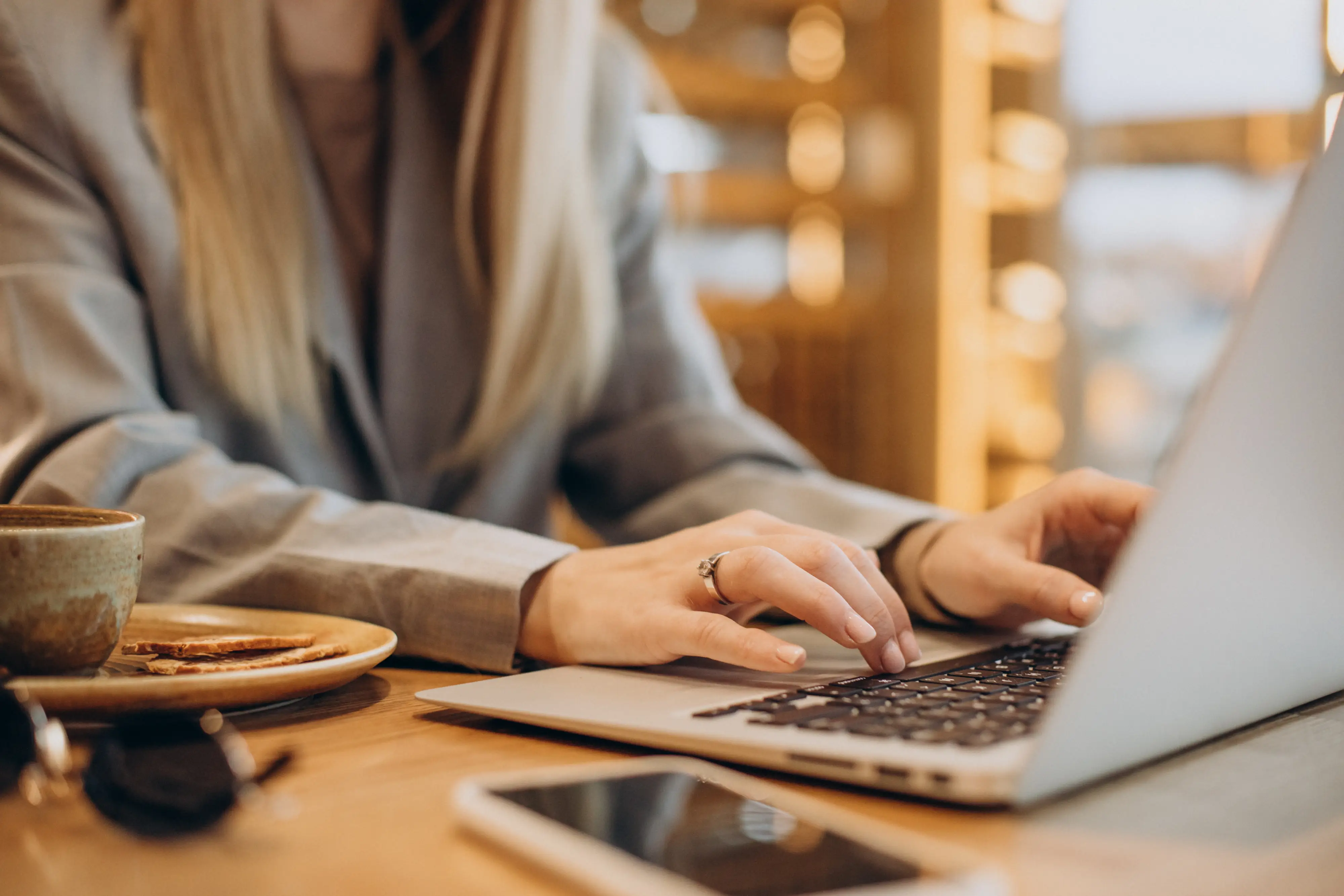 Woman with long blond hair typing on laptop keyboard at a wooden table in a cafe.