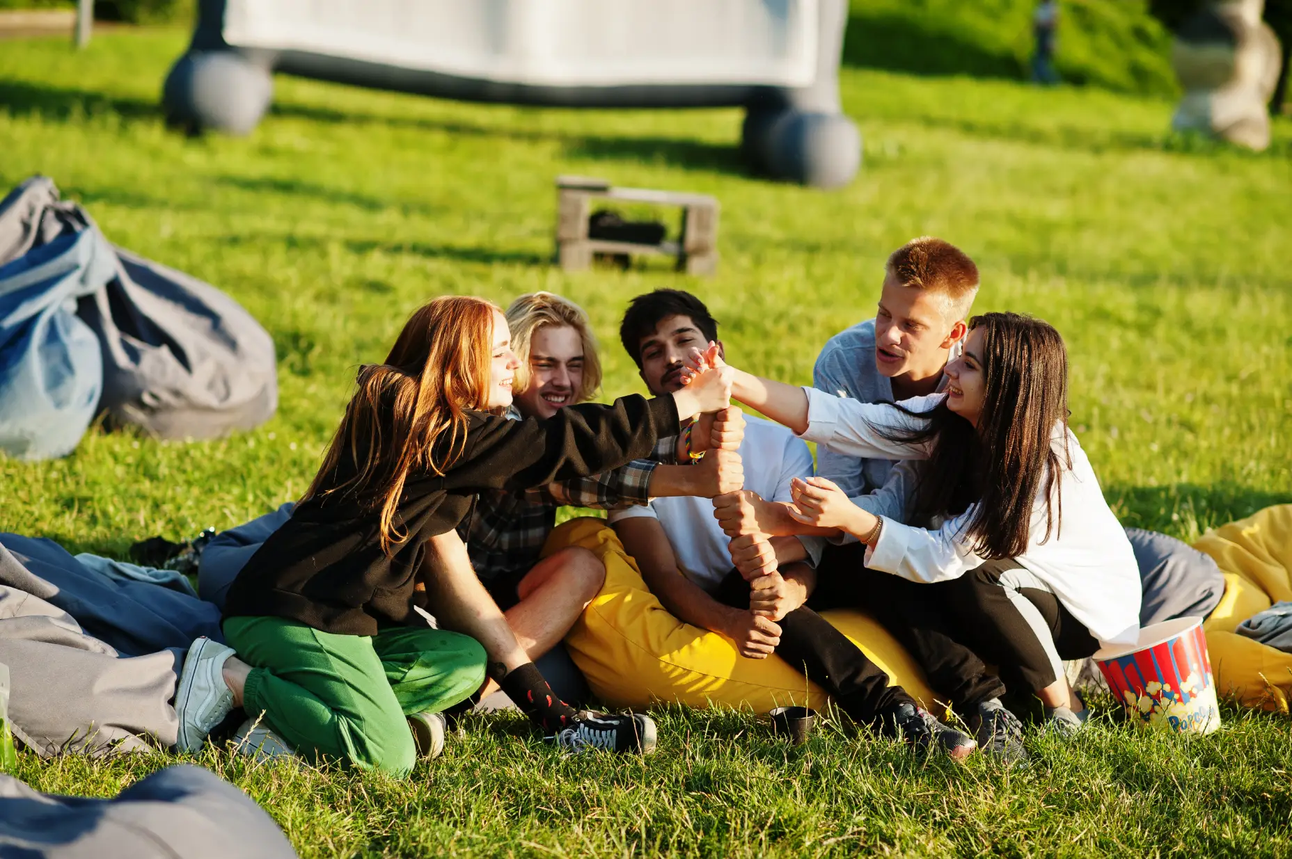 Group of young people sitting on grass outdoors, giving thumbs up and smiling, with popcorn nearby.