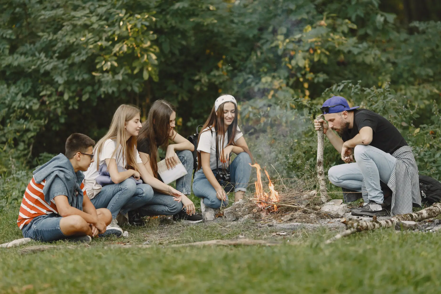 Group of five young people sitting outdoors around a small campfire in a forest setting.