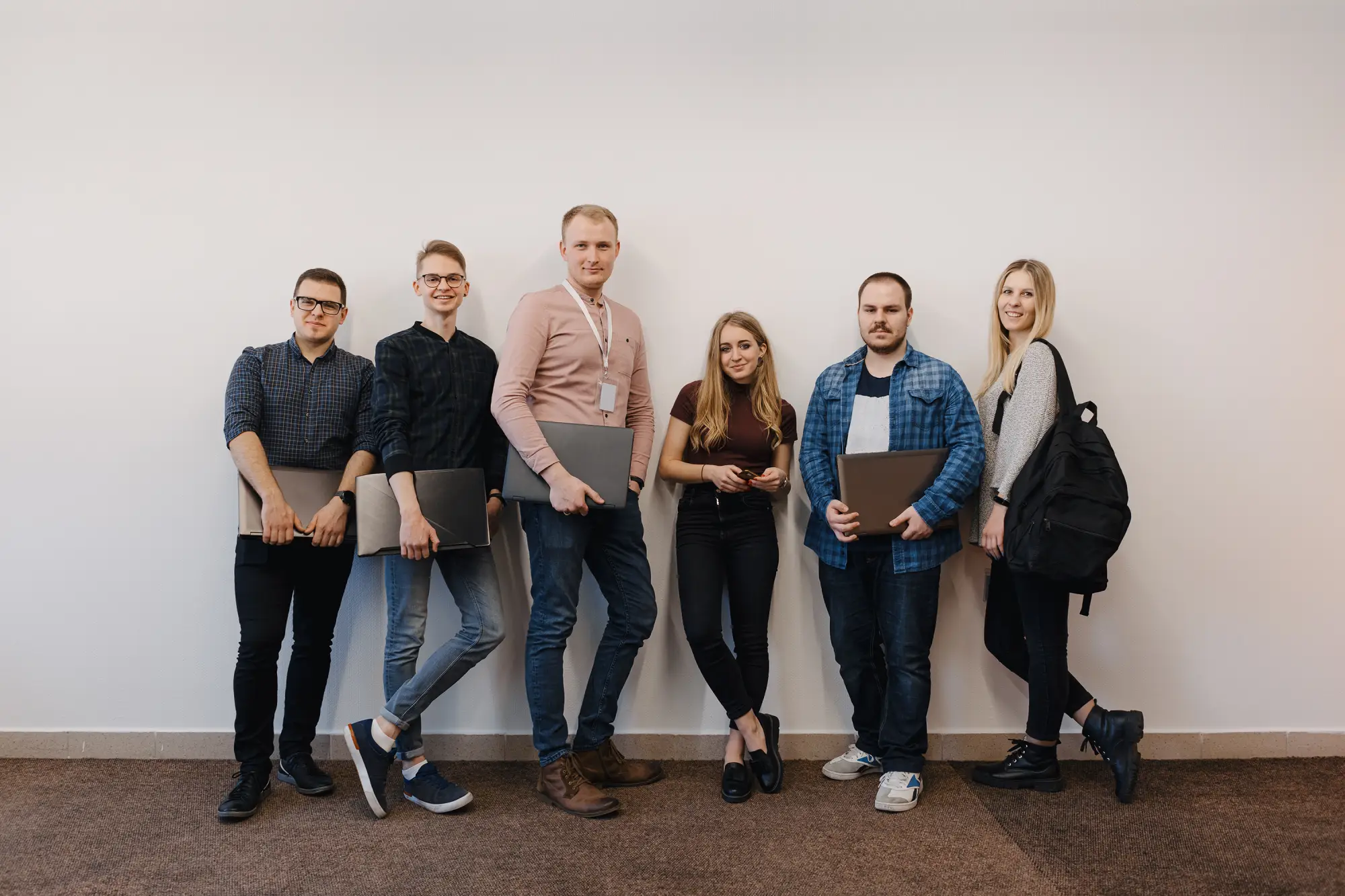 Group of six young adults standing against a wall, holding laptops and a backpack, casually dressed.