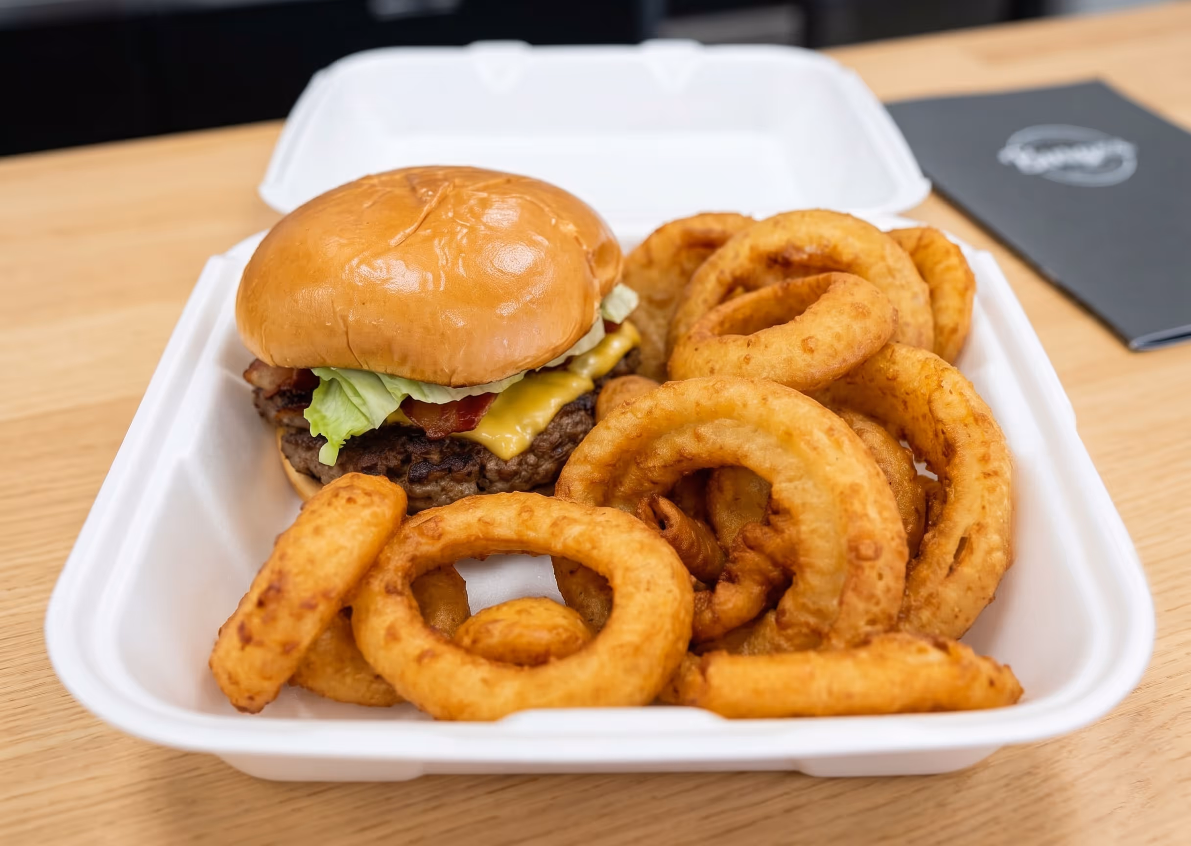 Cheeseburger with lettuce and bacon served with crispy onion rings in a white takeout container.