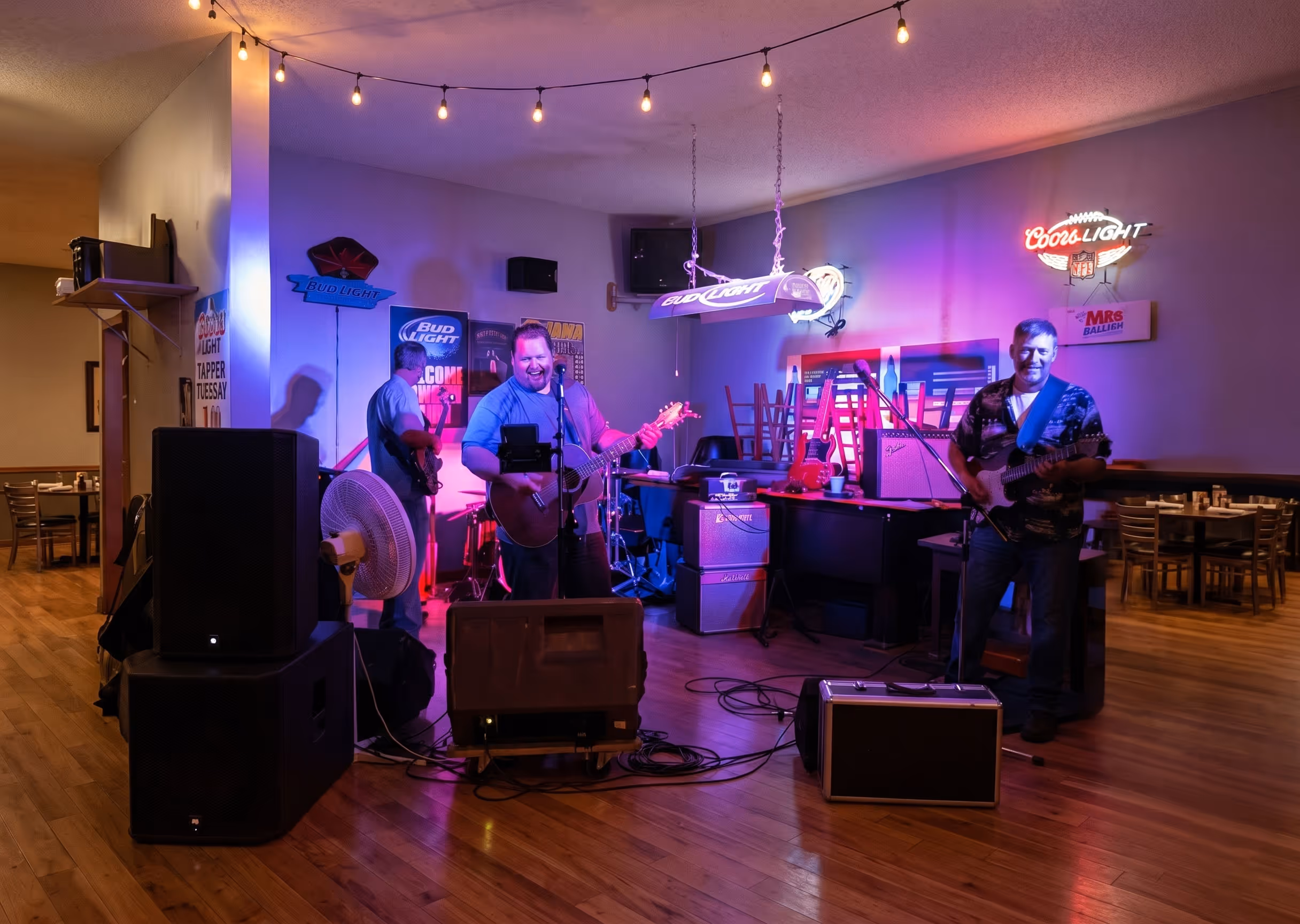 Three men are playing guitars and singing in a bar with purple lighting and music equipment.