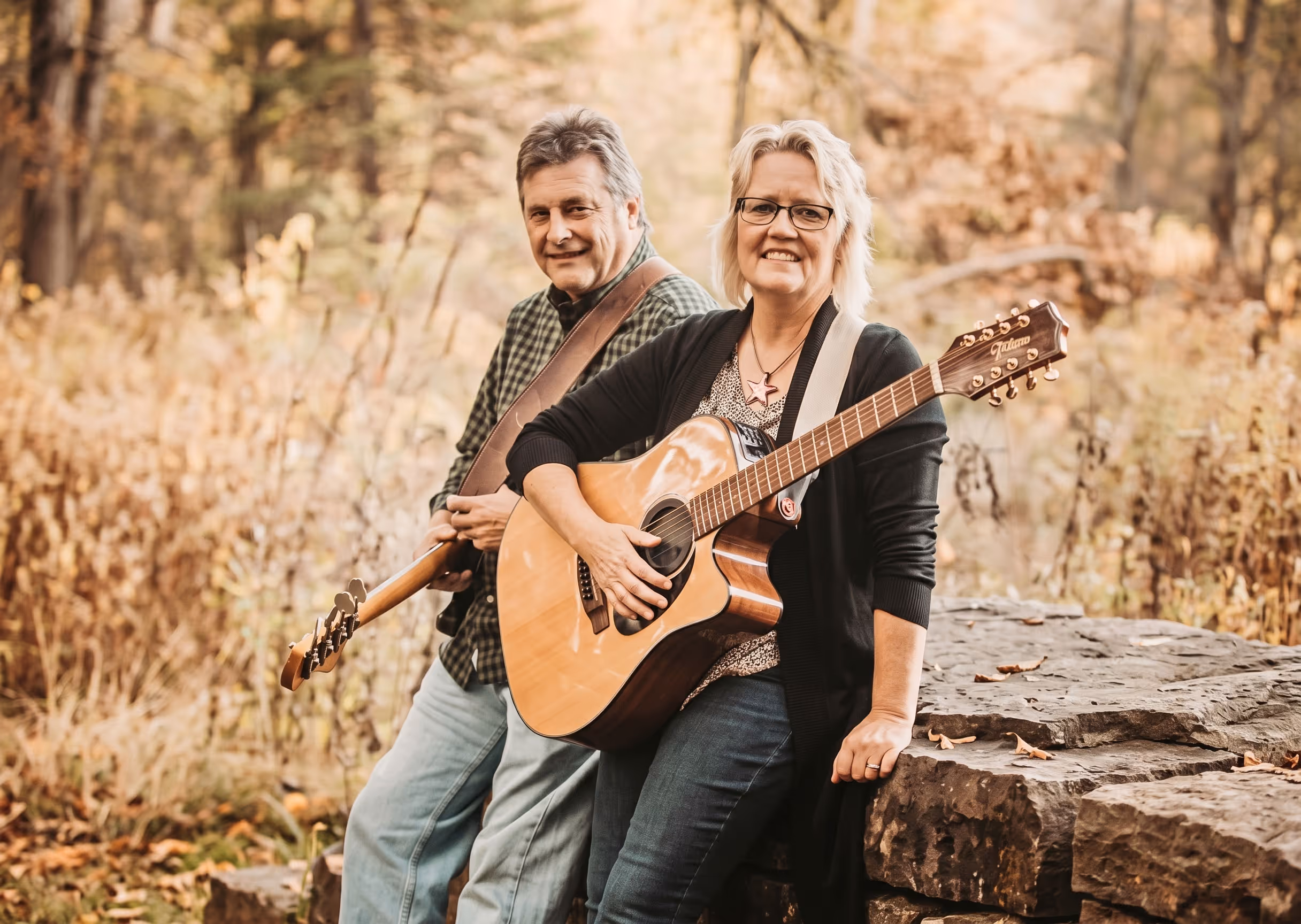 A smiling man and woman outdoors with acoustic guitars sitting on a stone ledge in autumn.