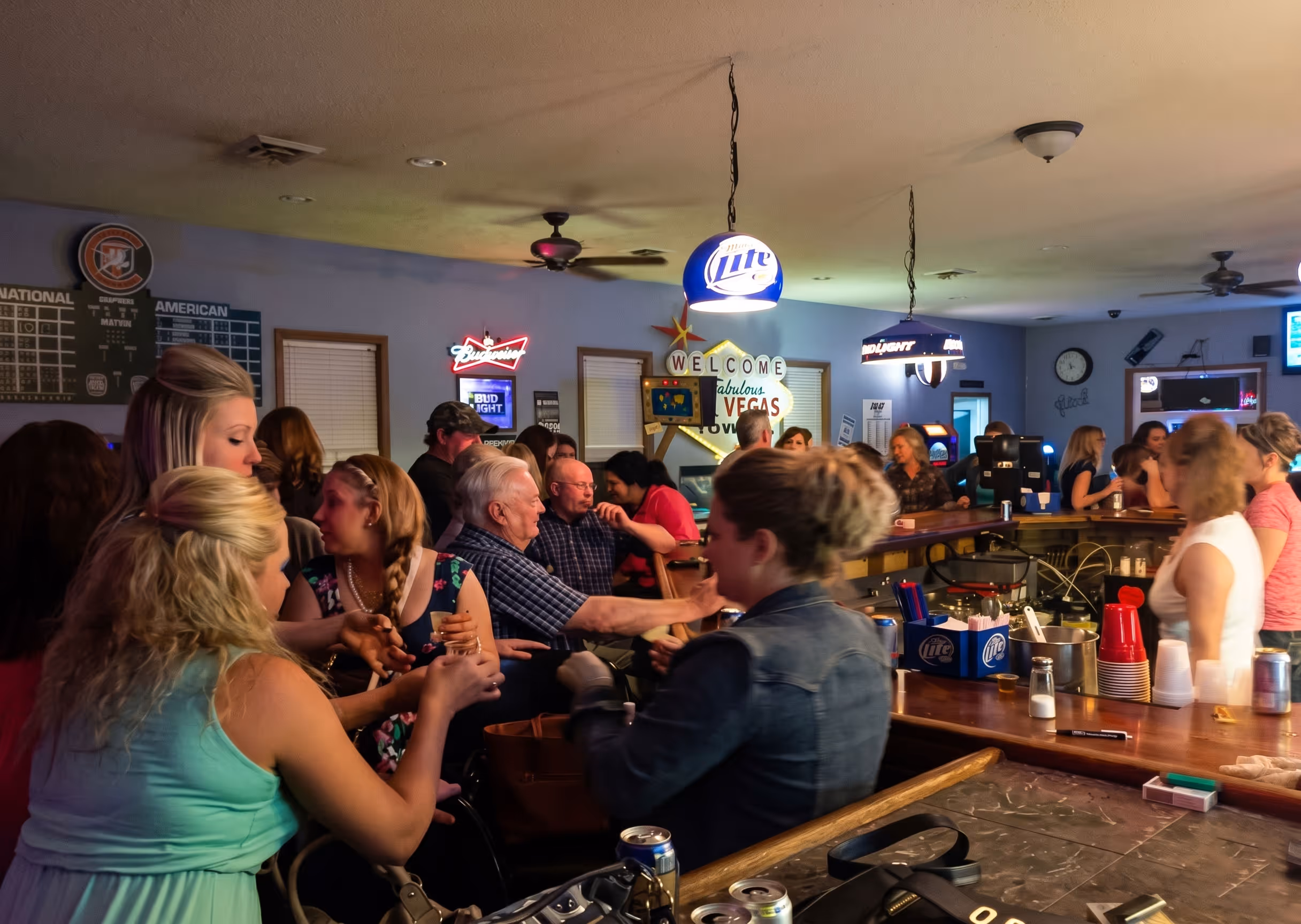 Crowded bar scene with people socializing near a wooden counter.