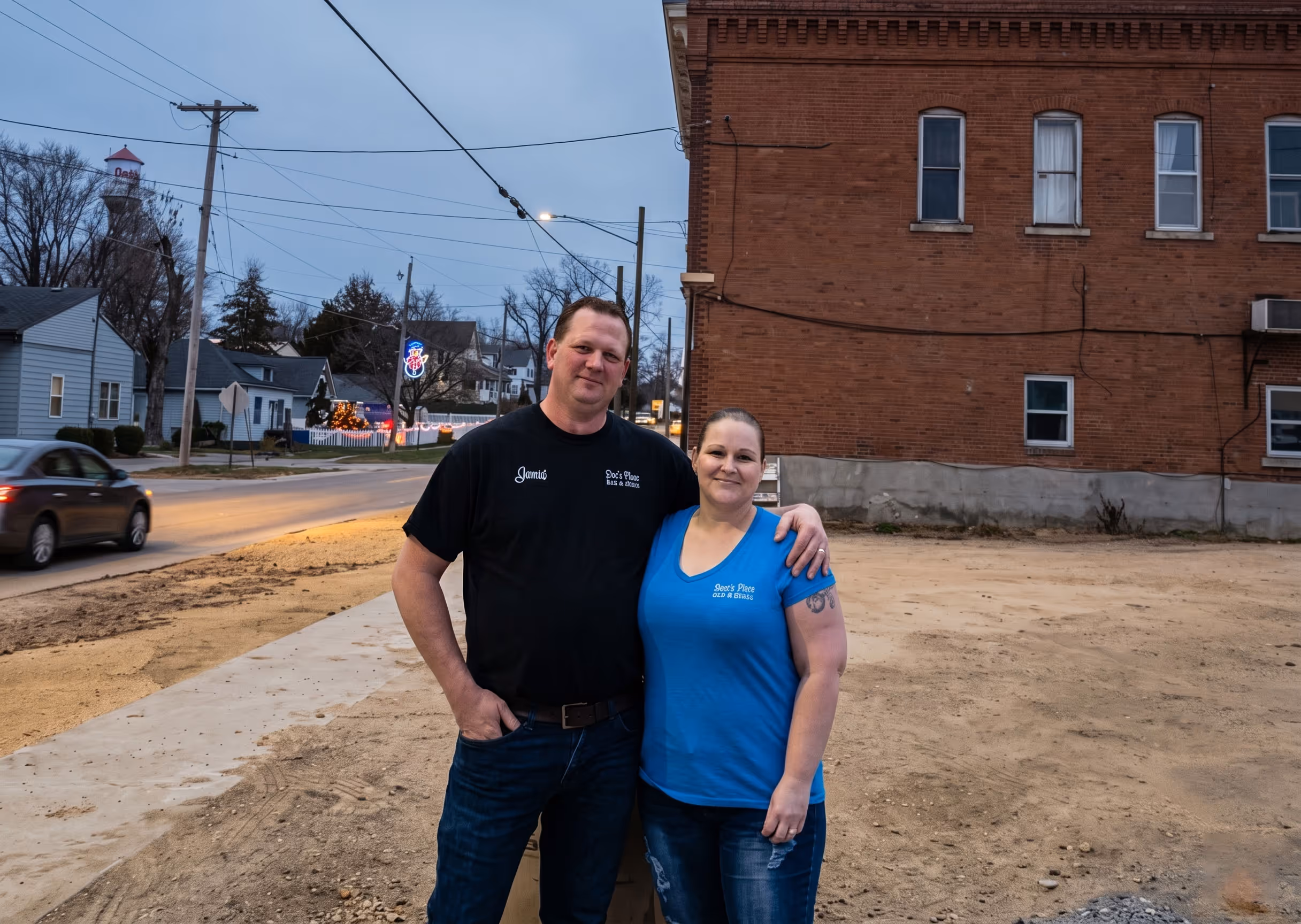 Man and woman standing arm in arm on a street corner next to a brick building at dusk.