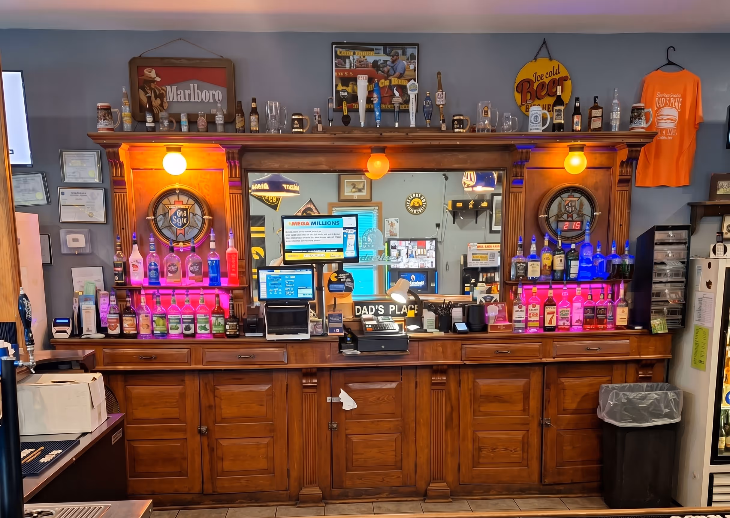 Wooden bar counter with colorful illuminated bottles, beer taps, a mirror, and a cash register.