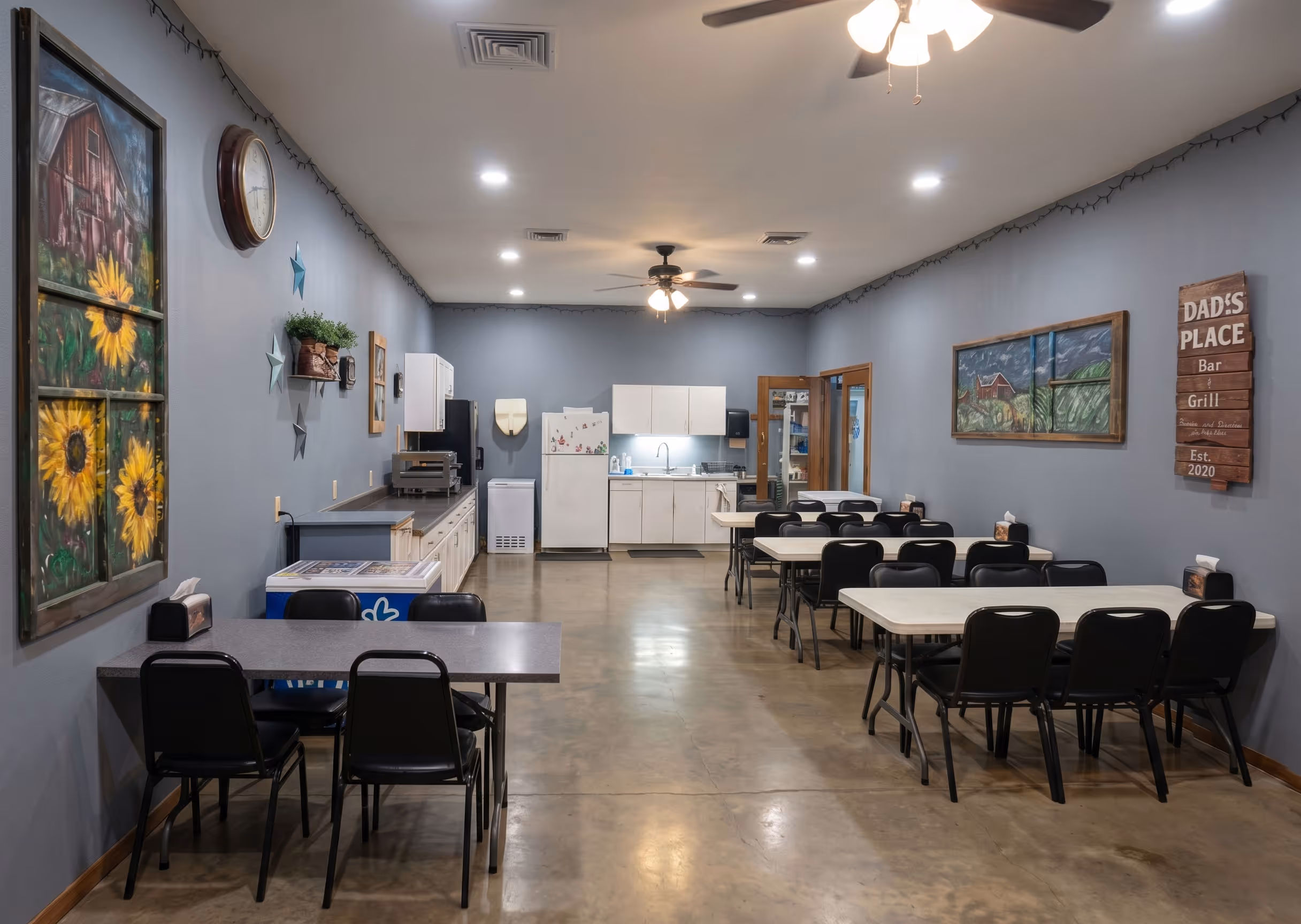 Room with gray walls, multiple tables with black chairs, a kitchen area, and decorative paintings.
