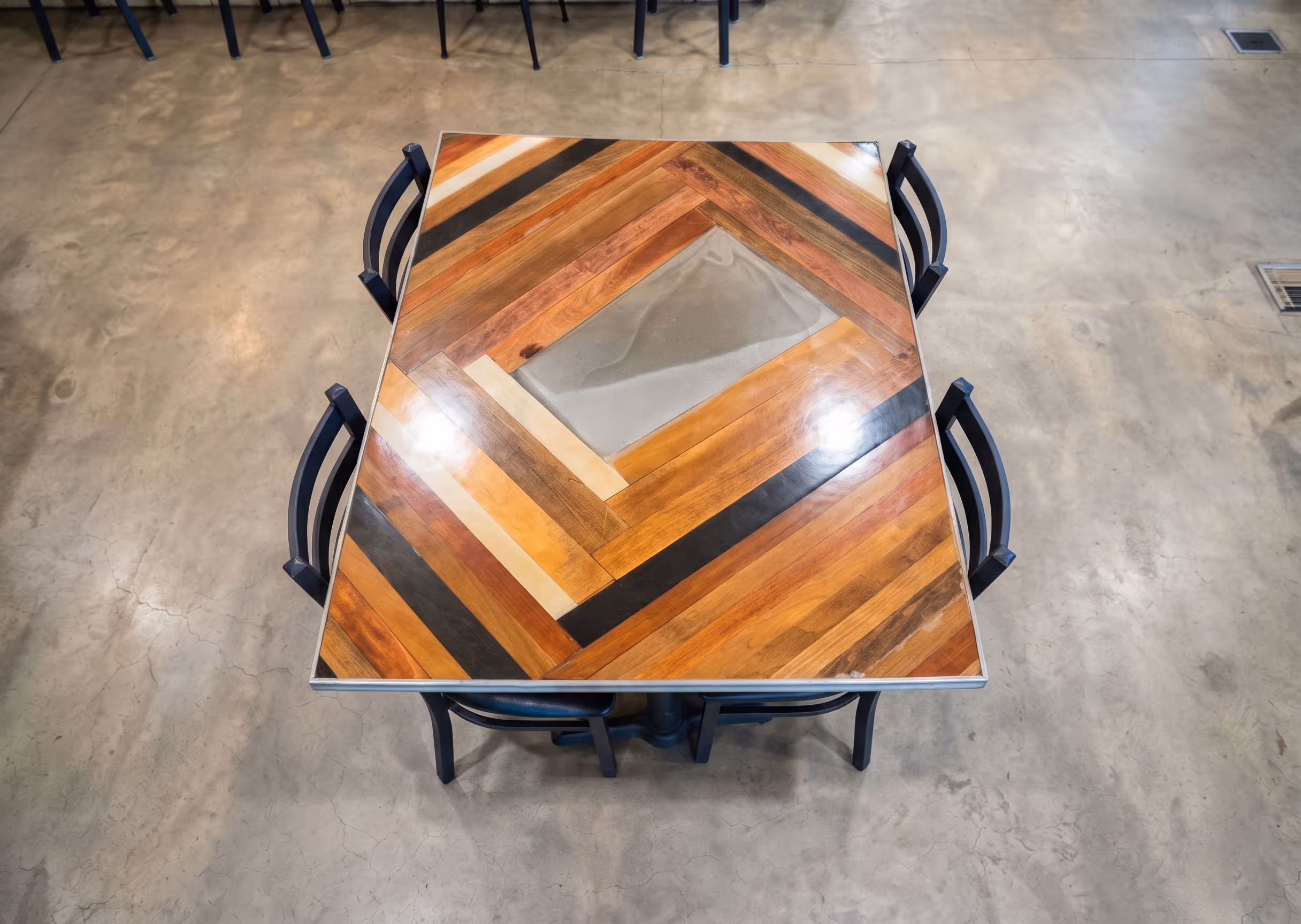 Square wooden table with a geometric pattern and four black chairs on a concrete floor.