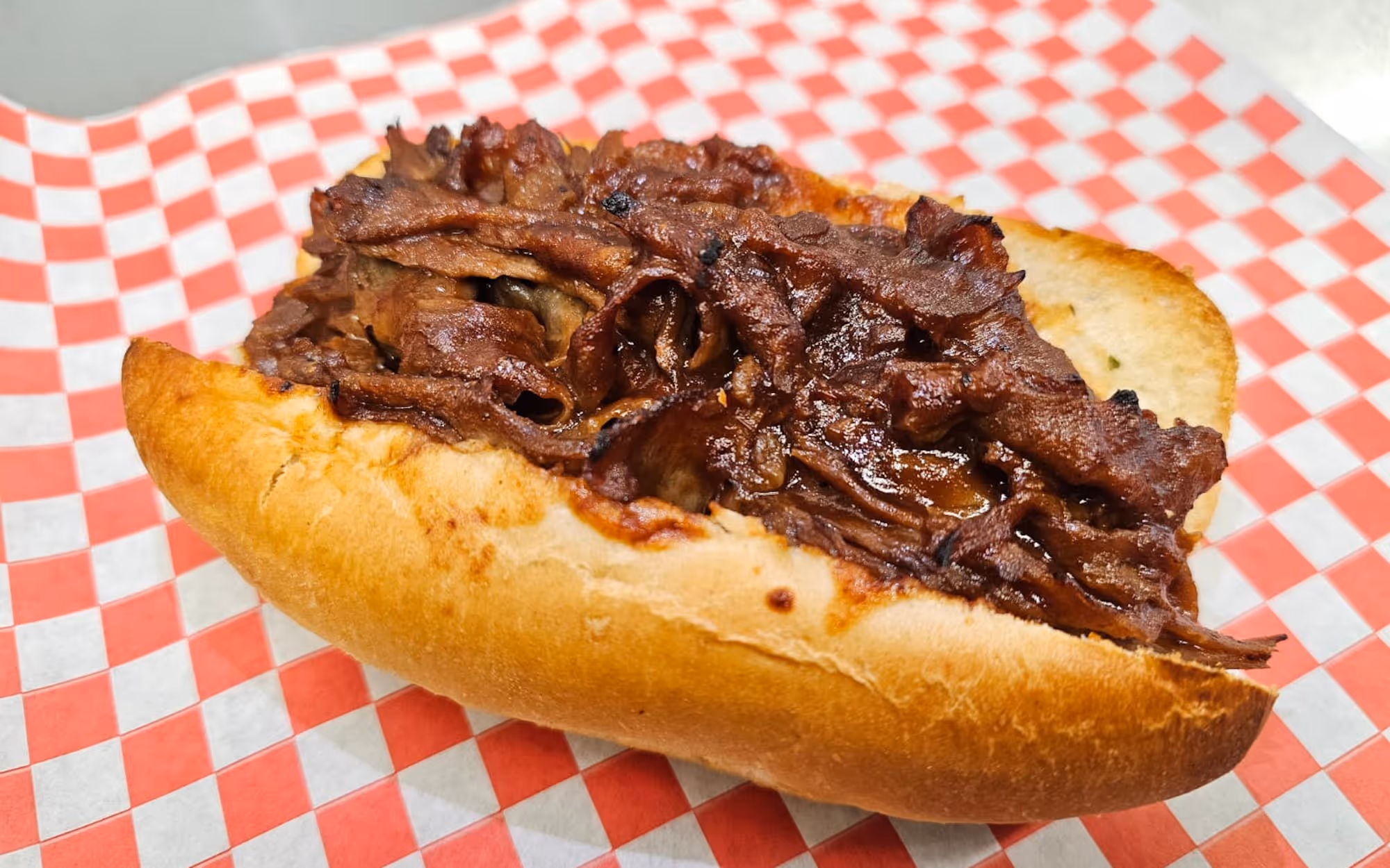 Steak sandwich with thinly sliced ribeye beef on a toasted hoagie roll atop red and white checkered deli paper.