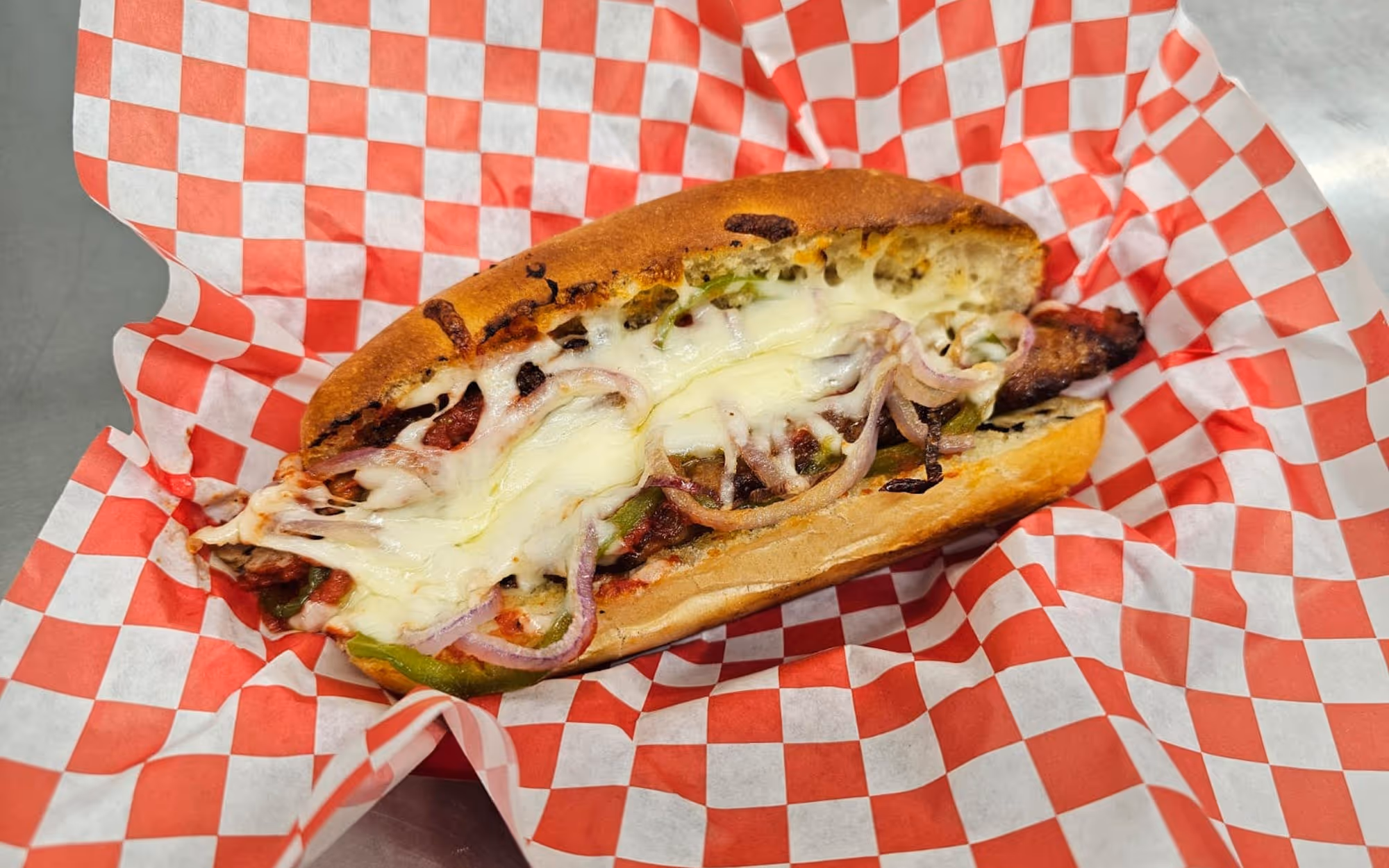 Philly cheesesteak sandwich with melted provolone cheese, onions, and peppers on a toasted roll over red checkered paper.