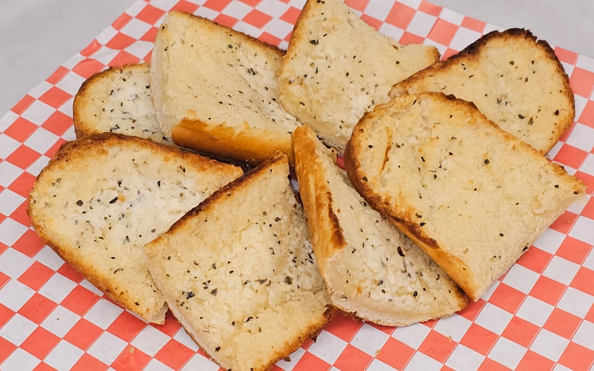 Toasted garlic bread slices with dried herbs on a red and white checkered deli paper.