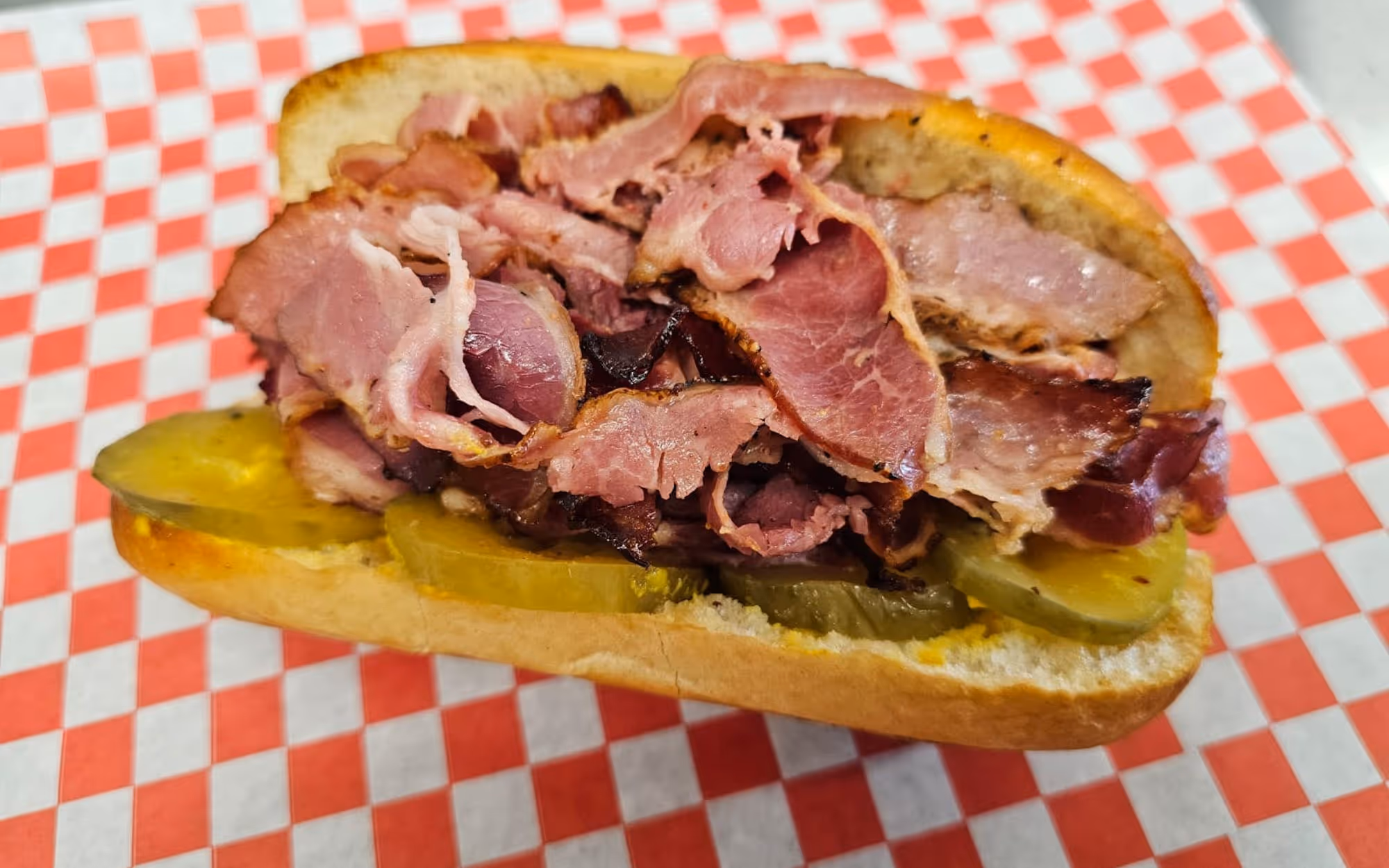 Hot pastrami sandwich with sliced pickles on a toasted roll over a red and white checkered deli liner.