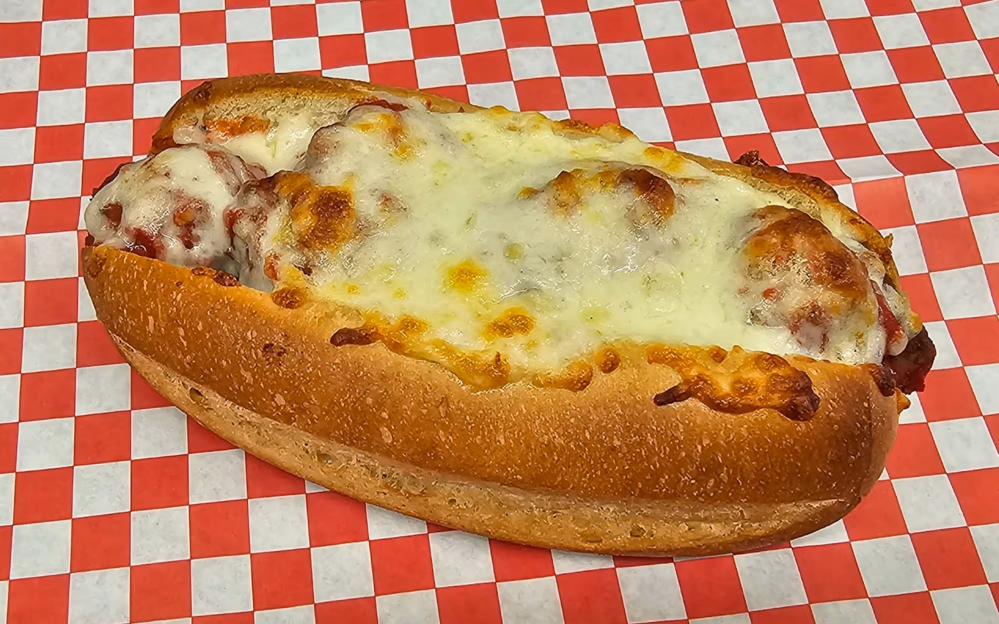 A meatball sub sandwich with melted mozzarella cheese and marinara sauce on a toasted hoagie roll over red checkered paper.