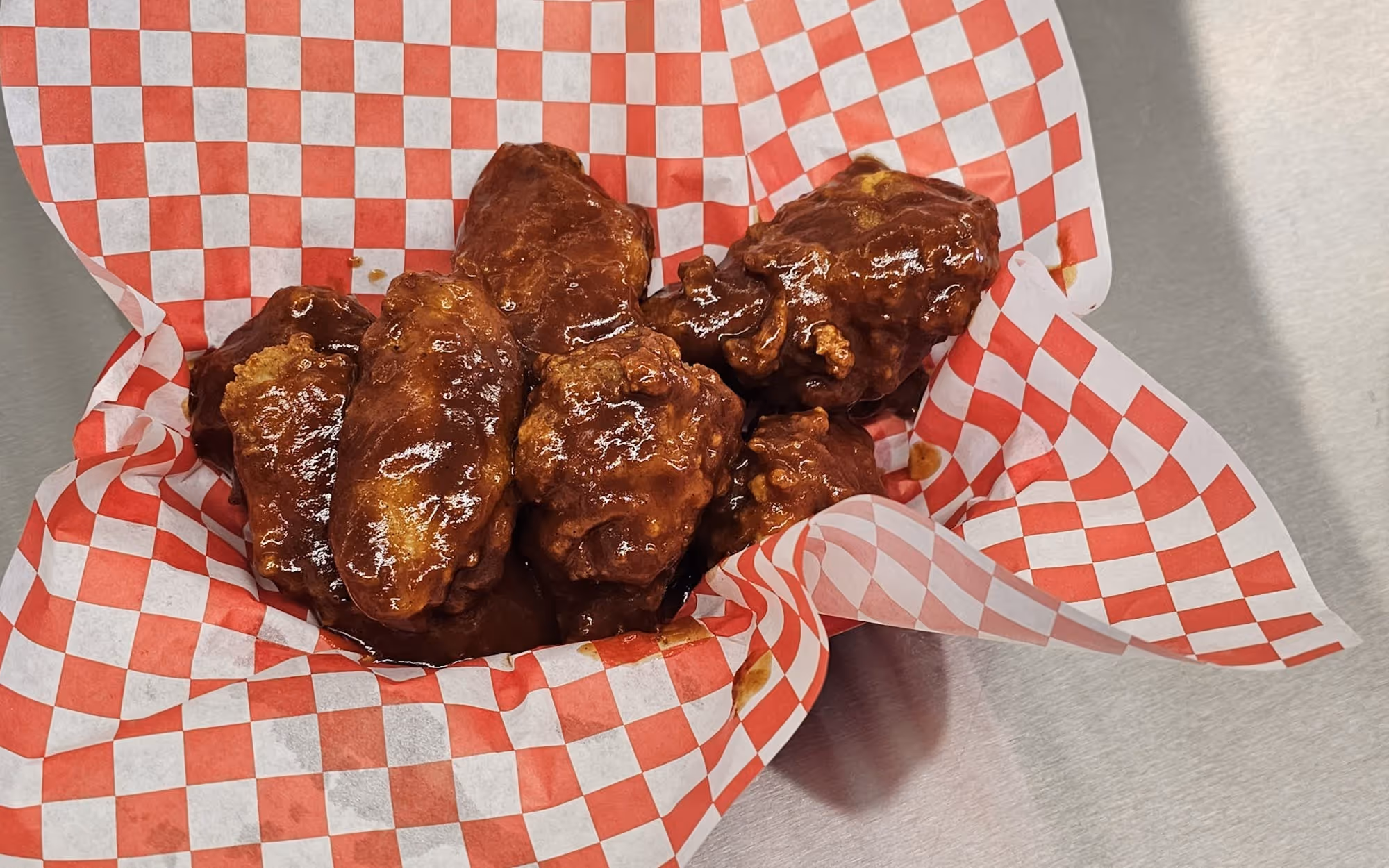 Crispy chicken wings coated in thick red barbecue sauce served on red and white checkered deli paper.