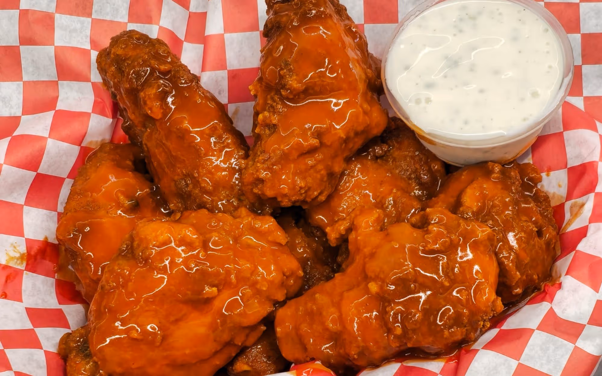 Crispy buffalo chicken wings coated in spicy orange sauce with a side of ranch dressing in a red checkered basket liner.