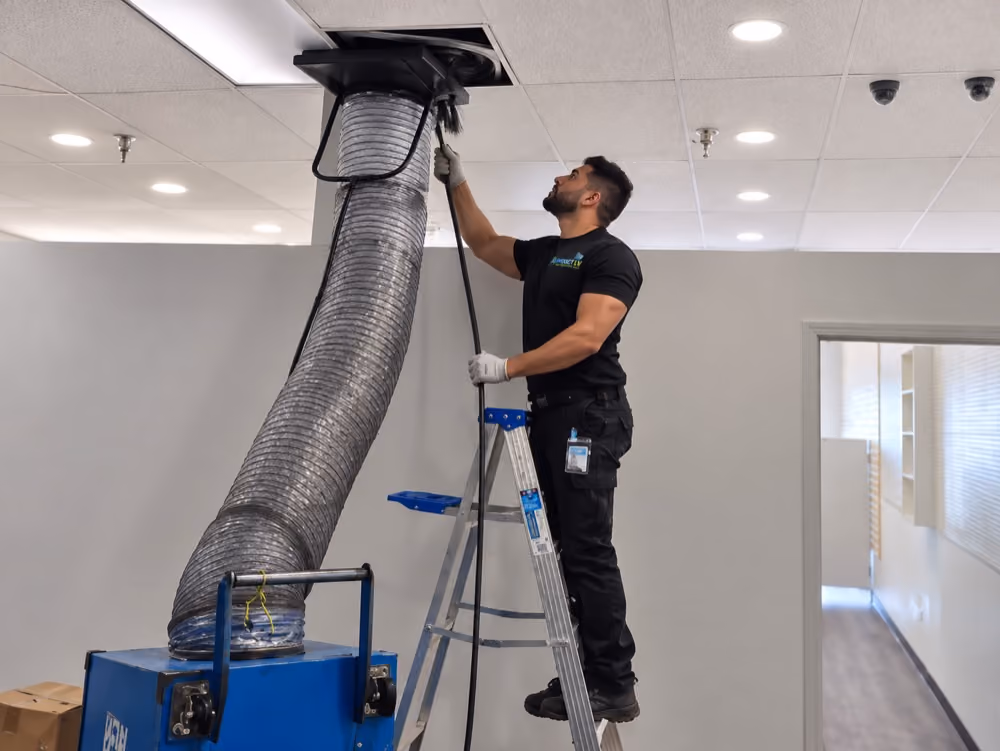 Air Duct LV technician on a ladder connecting a flexible hose to a ceiling vent.