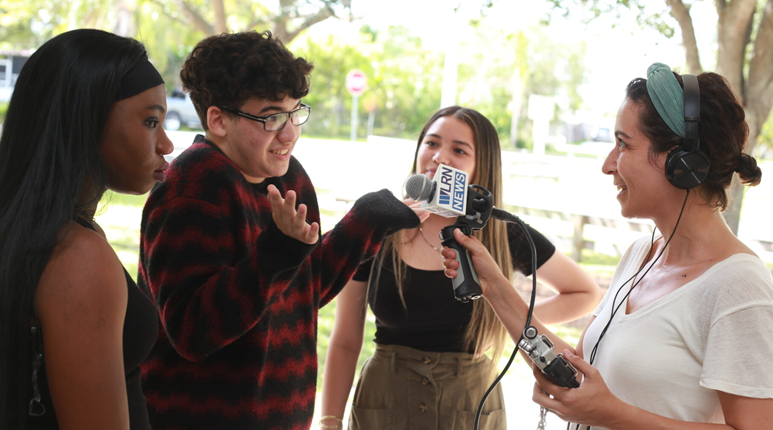 A young man speaks into a microphone held by a woman wearing headphones while two other young people stand nearby outdoors.