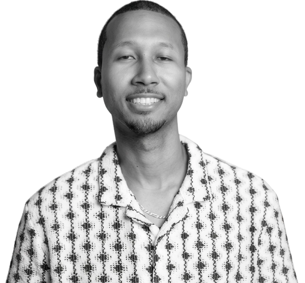 Black-and-white portrait of a smiling man wearing a patterned shirt and chain necklace against a bright yellow background.