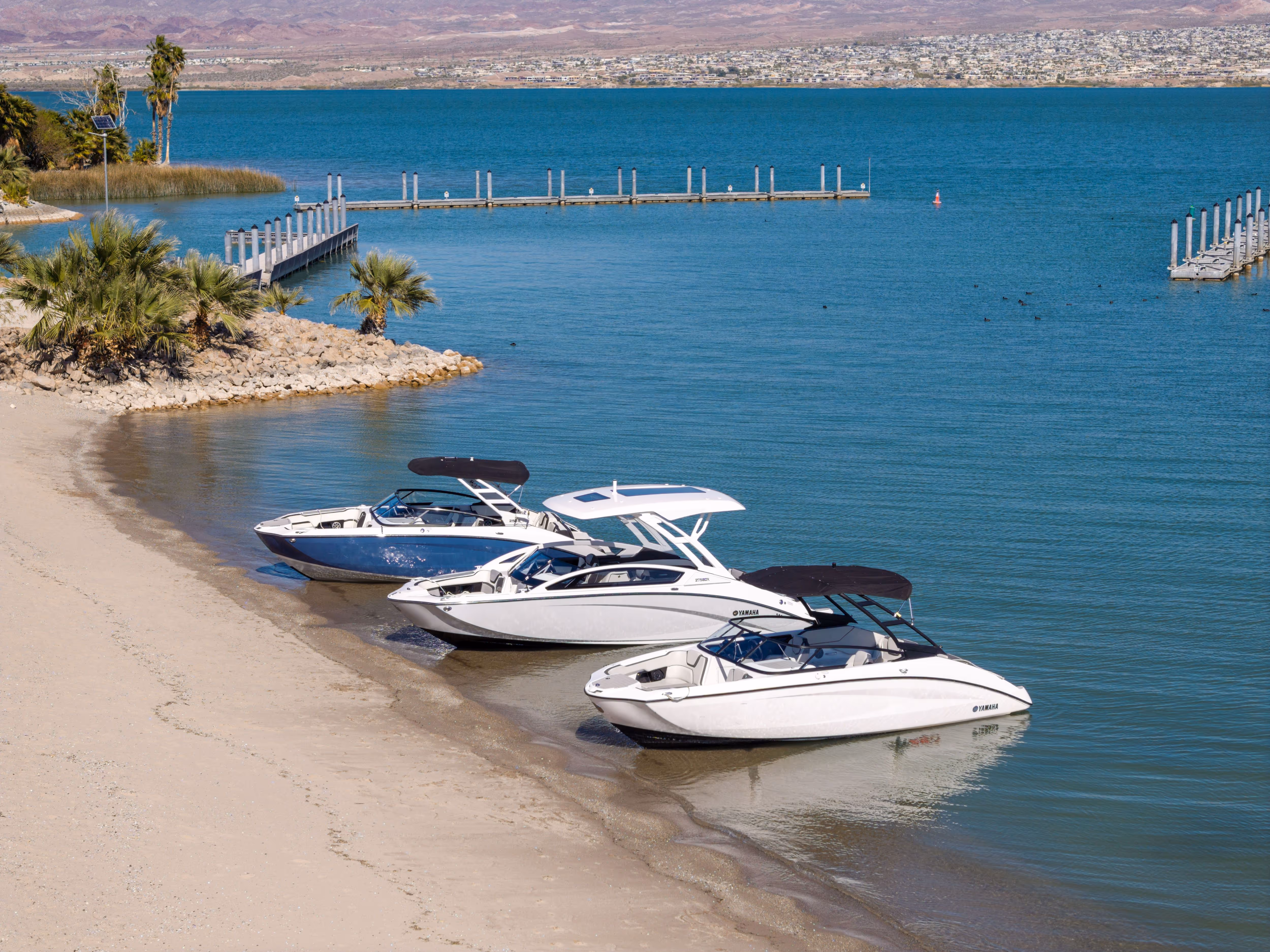 Three motorboats parked on a sandy beach with blue water and docks in the background.