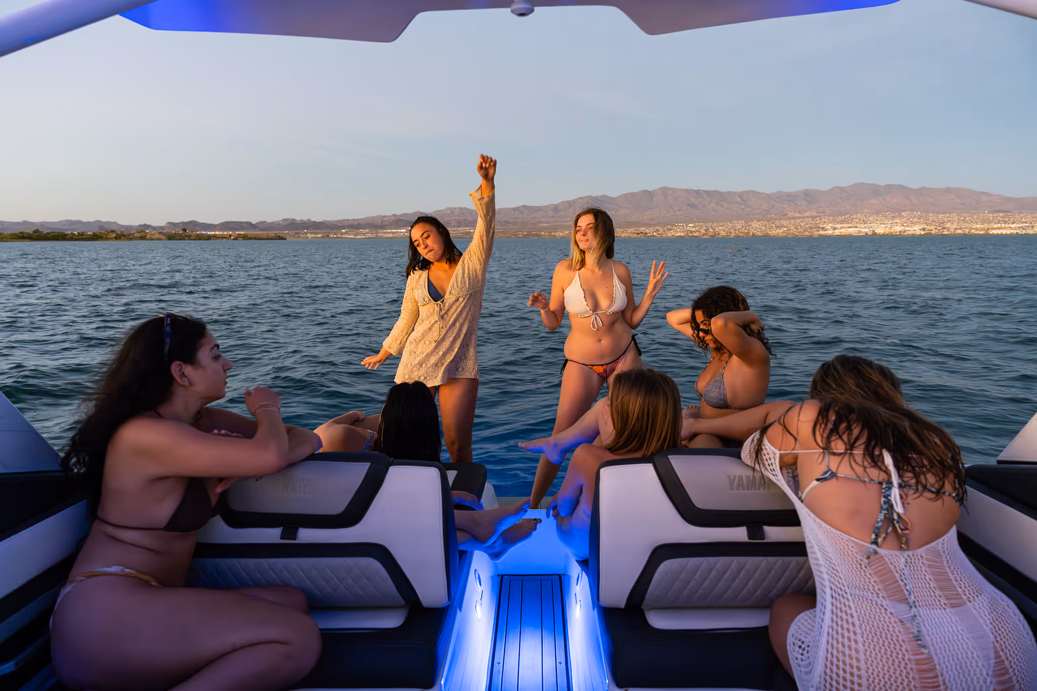 Group of young women in swimsuits on a boat with ocean and mountains in the background at sunset.