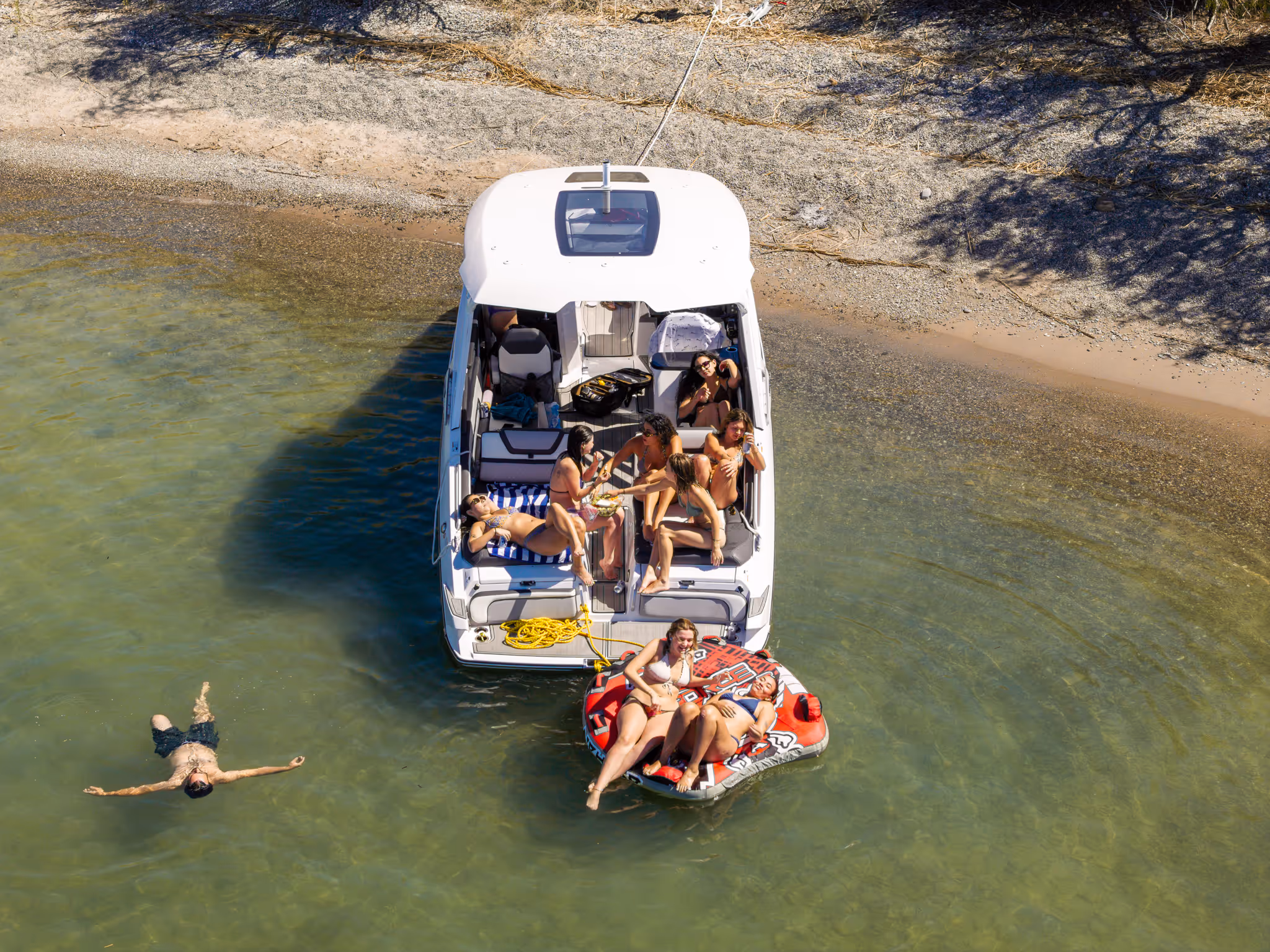 People relaxing on a boat and inflatable tube near a sandy shore in shallow green water.