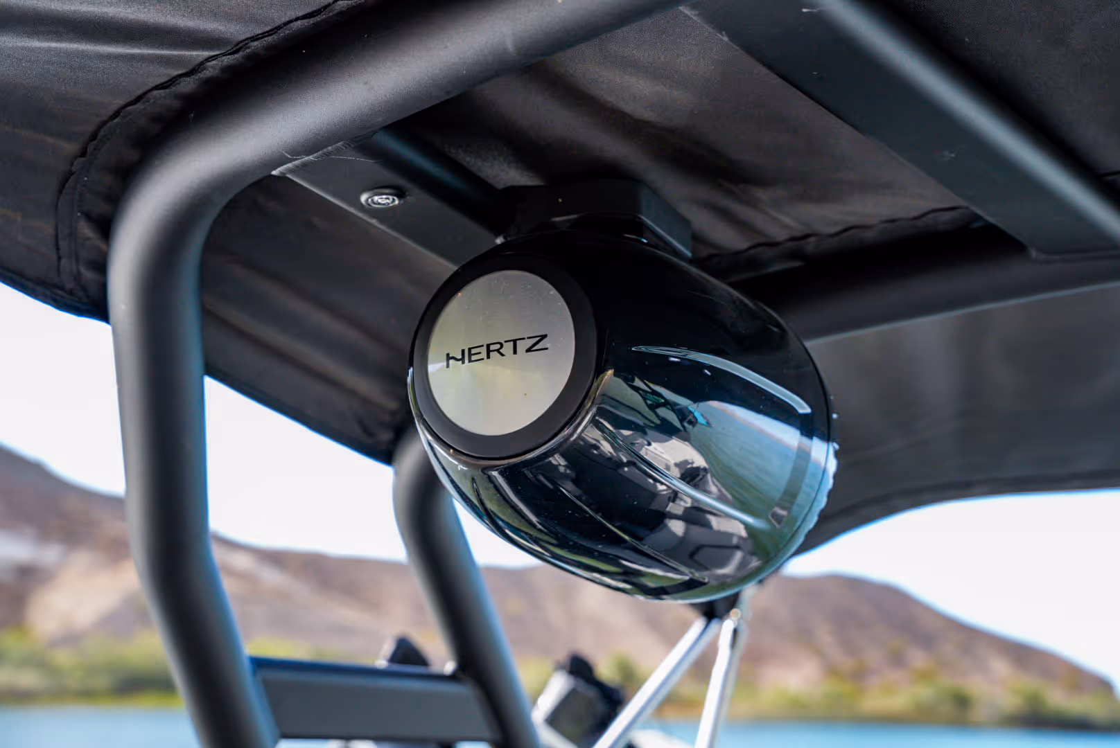 Black Hertz marine speaker mounted under a boat canopy with water and hills in the background.