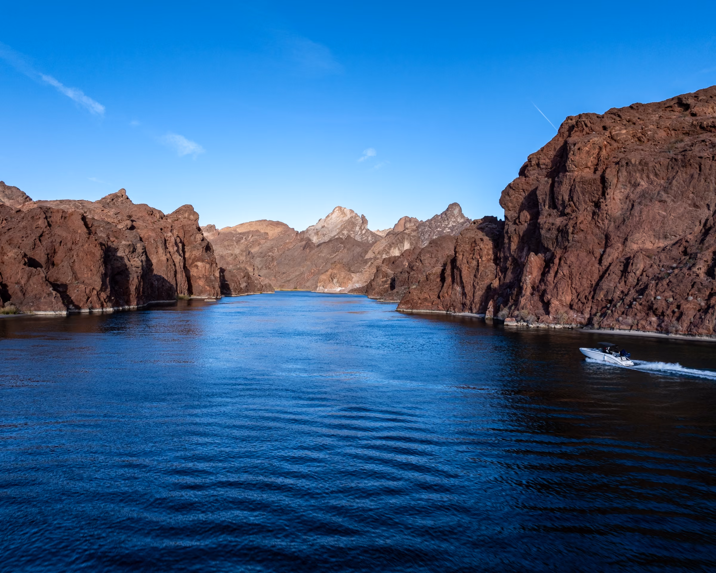A motorboat speeding on a wide river surrounded by tall, rocky brown cliffs under a clear blue sky.