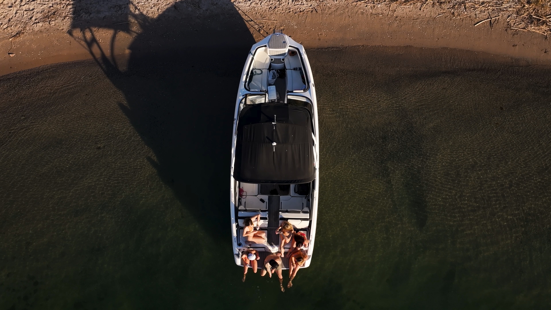 Overhead view of a boat with six people sitting at the rear near shallow water and a sandy shore.