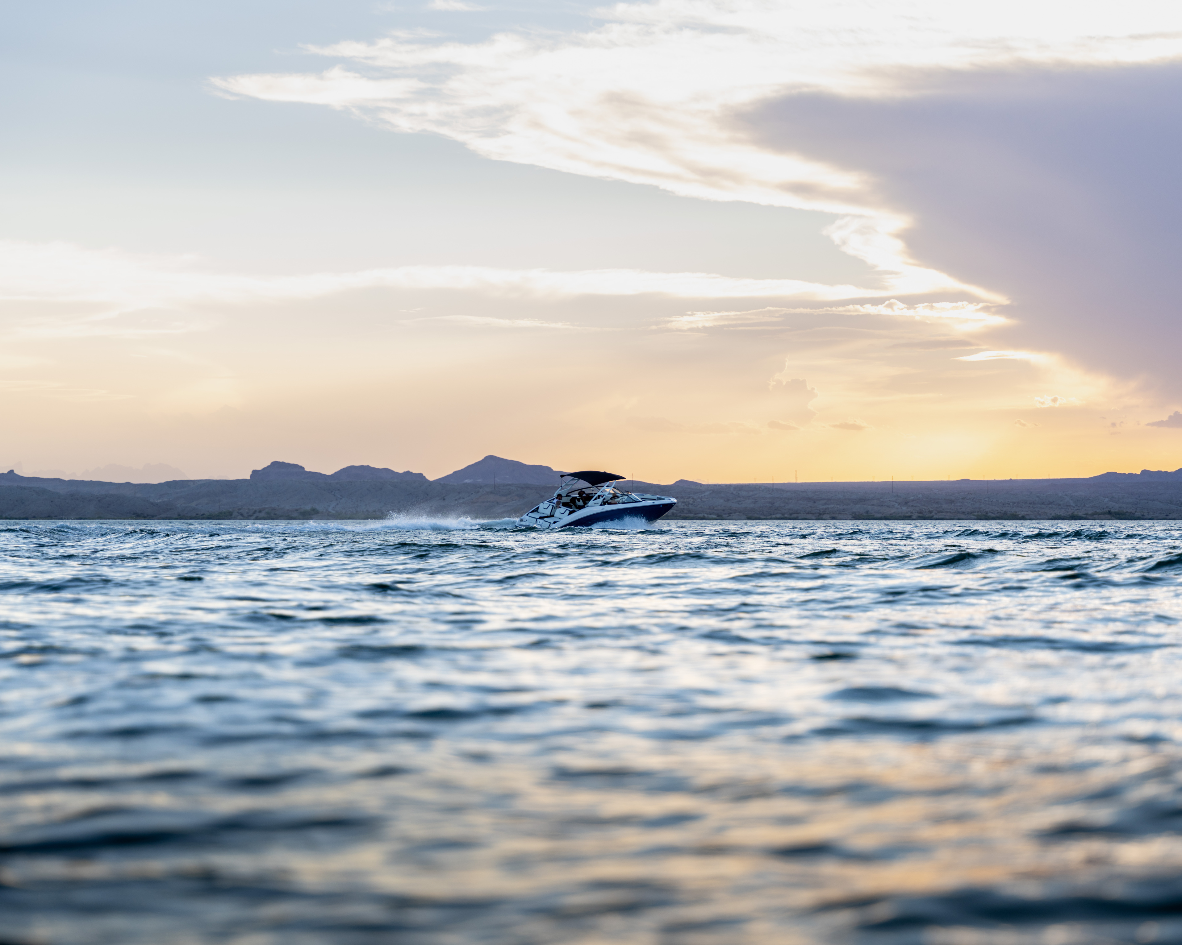 Speedboat moving on calm water with mountainous shore and sunset sky in the background.