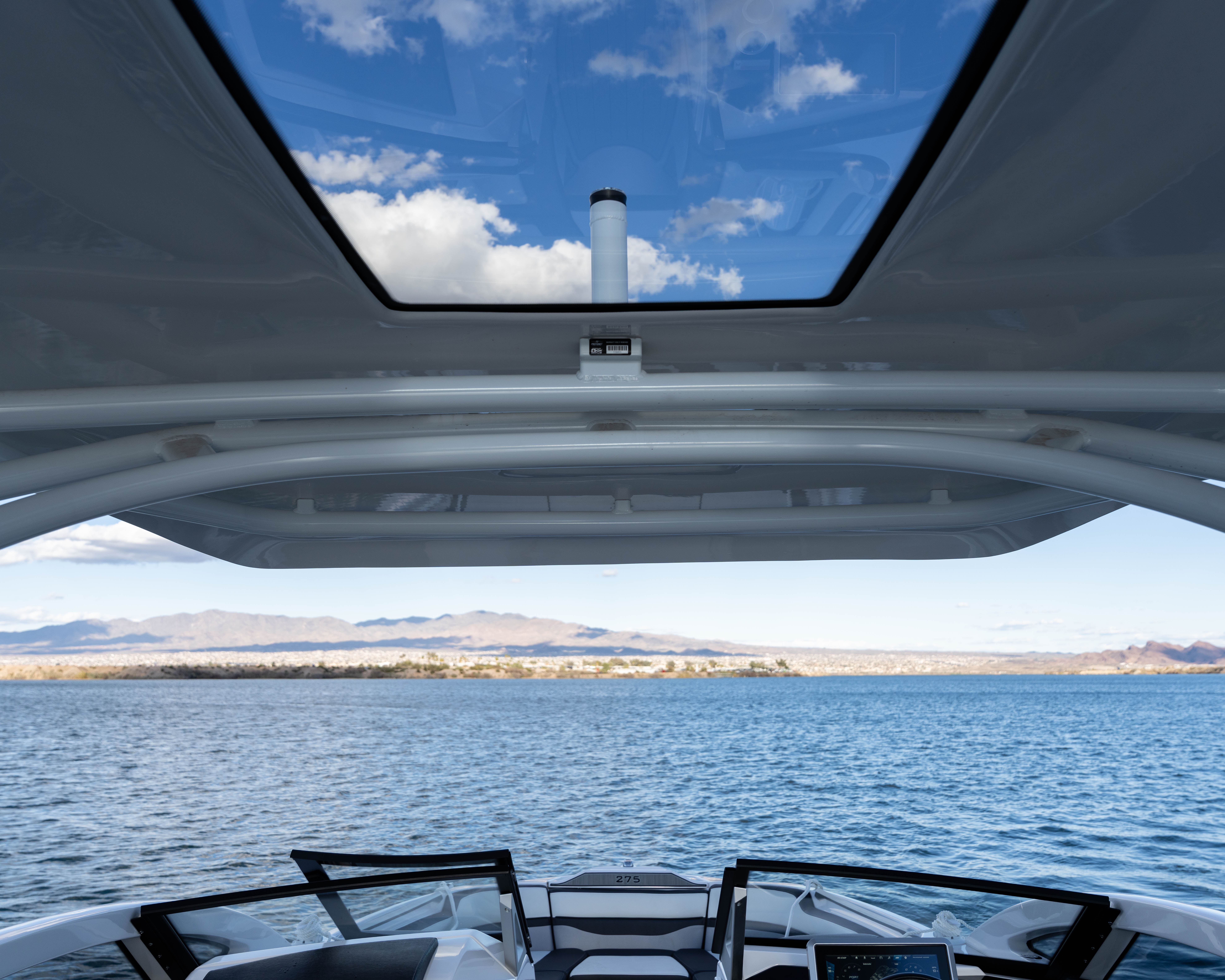 View from a boat cockpit showing water, distant shore, mountains, and clouds through a roof window.