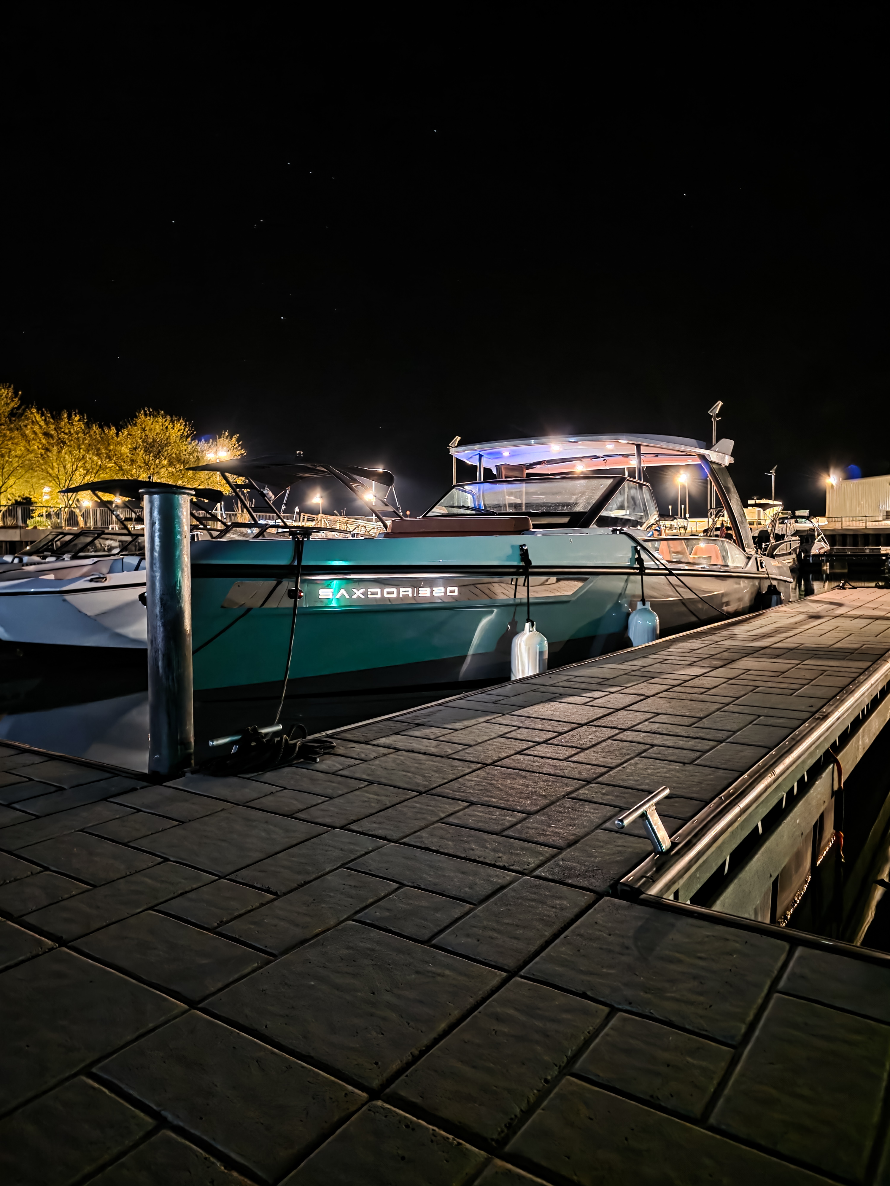 Teal motorboat docked at a marina at night with illuminated trees and other boats in the background.