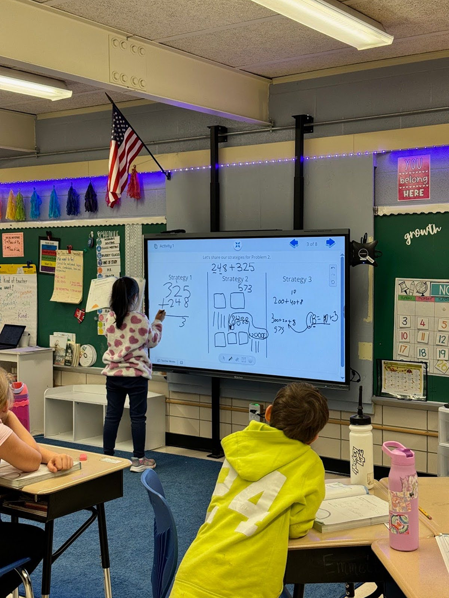 Child in a classroom solving math problems on a digital board with students watching.