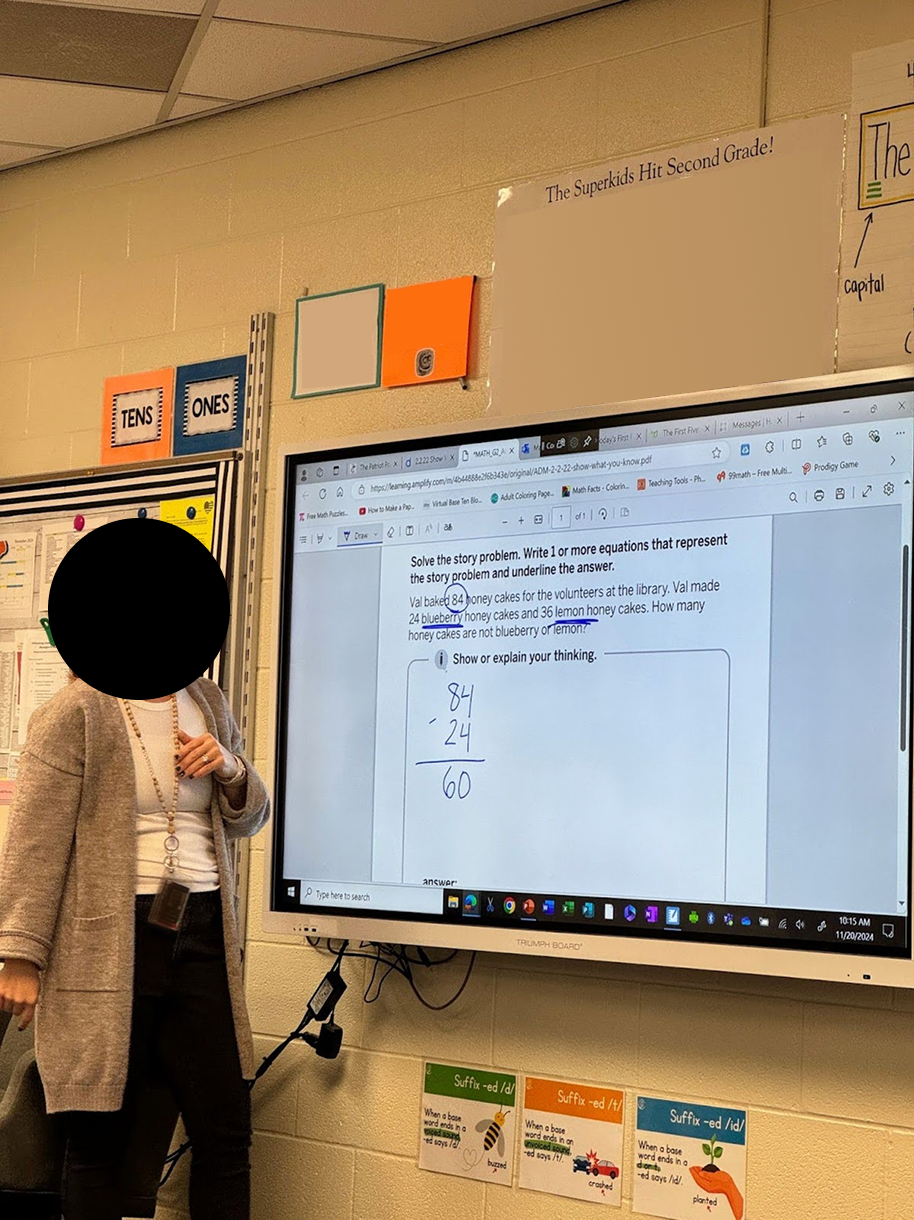 Teacher standing beside a screen showing a math word problem and subtraction solution in a classroom.