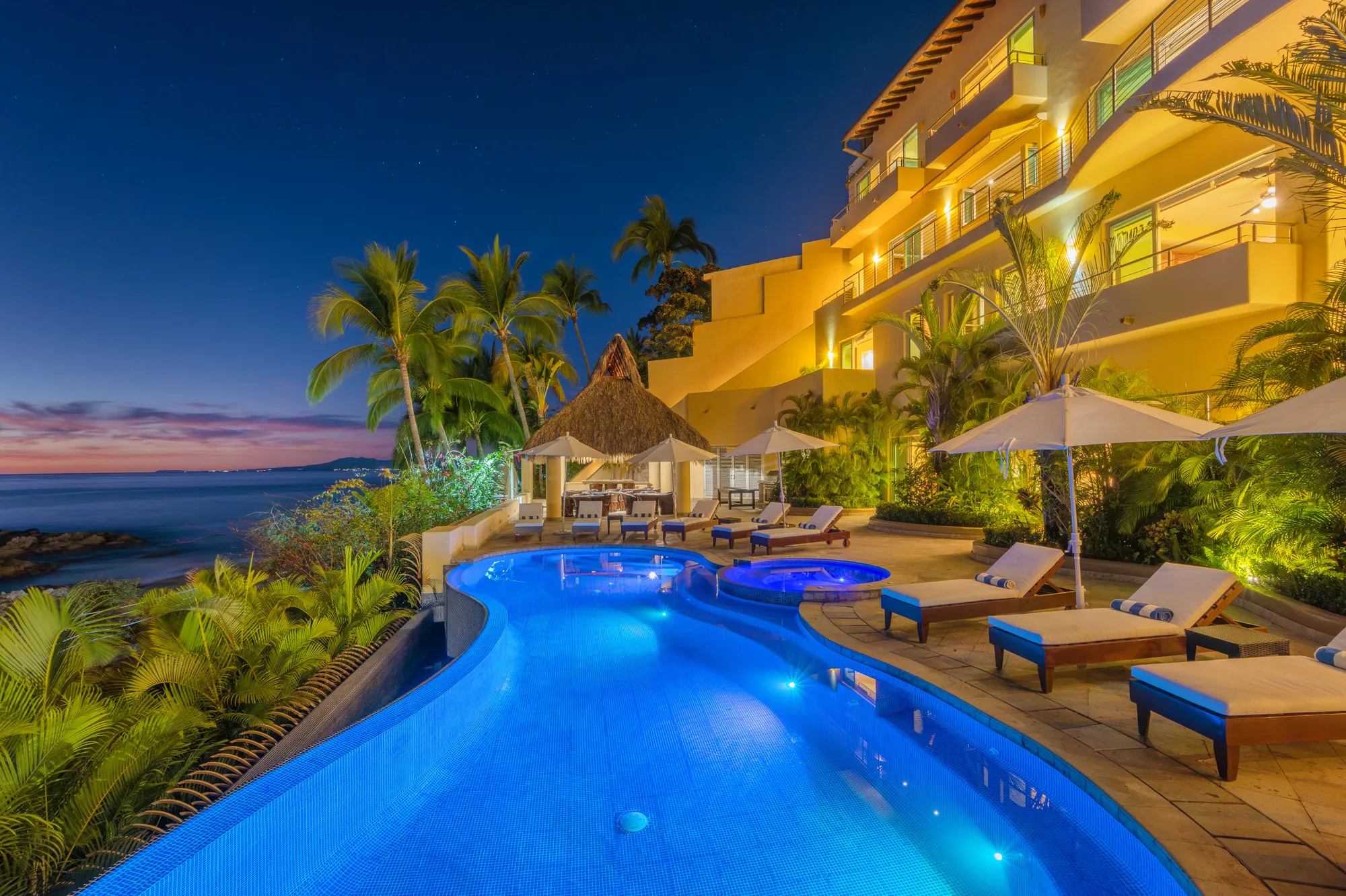Heated saltwater infinity pool and thatched-roof palapa bar at Villa Montecito illuminated at night, set against the four-level villa exterior and palm trees.