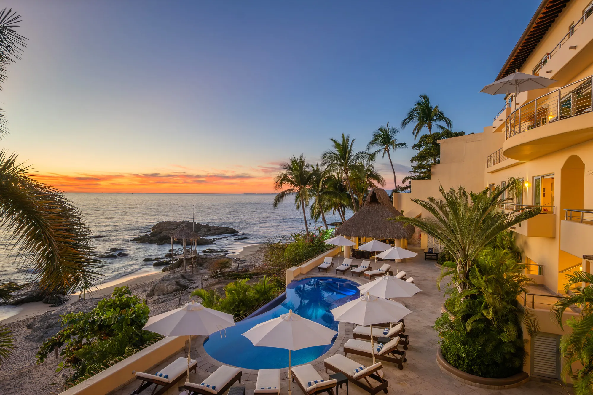 Sunset view across the upper terrace at Villa Montecito with palm trees, lounge chairs, and the heated infinity pool overlooking the Pacific Ocean in Conchas Chinas, Puerto Vallarta.