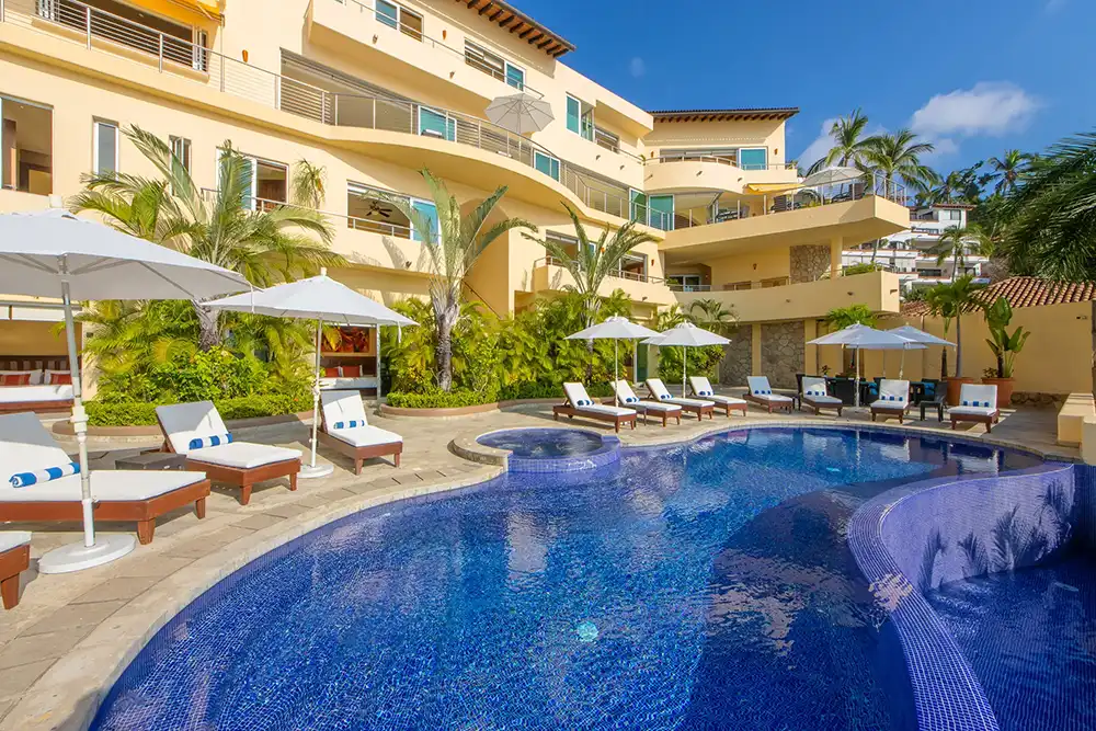 Outdoor pool area with lounge chairs, umbrellas, tropical plants, and multi-story building under blue sky.