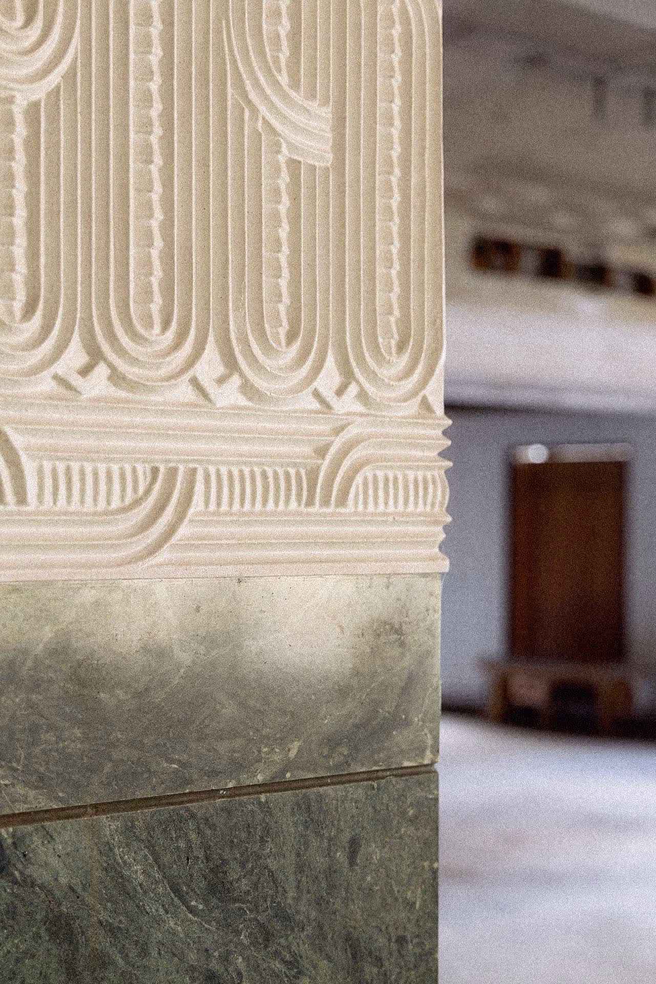 Close-up of ornate stone or plaster wall detailing with geometric and linear carved patterns in cream and greenish tones, with an out-of-focus interior space featuring a door and bench in the background.