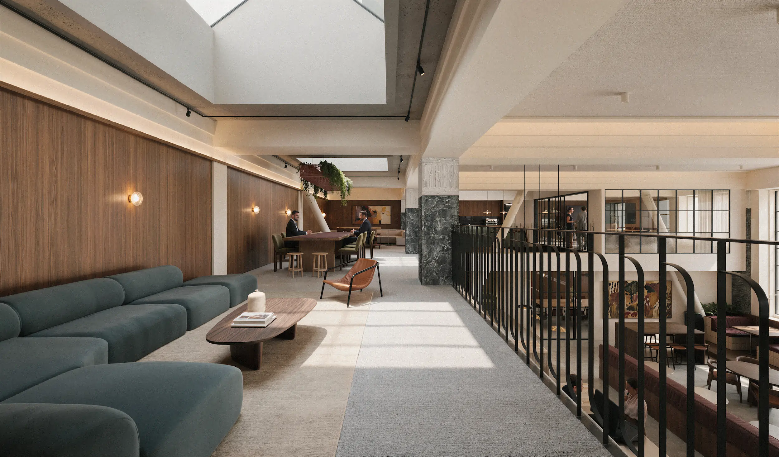 Modern coworking space with a large teal sectional sofa, a wooden coffee table with books and a plant, brown leather chair, and two men working at a wooden table. The area has wood-paneled walls, skylights, and black metal railing overlooking a lower floor with more seating and people working.