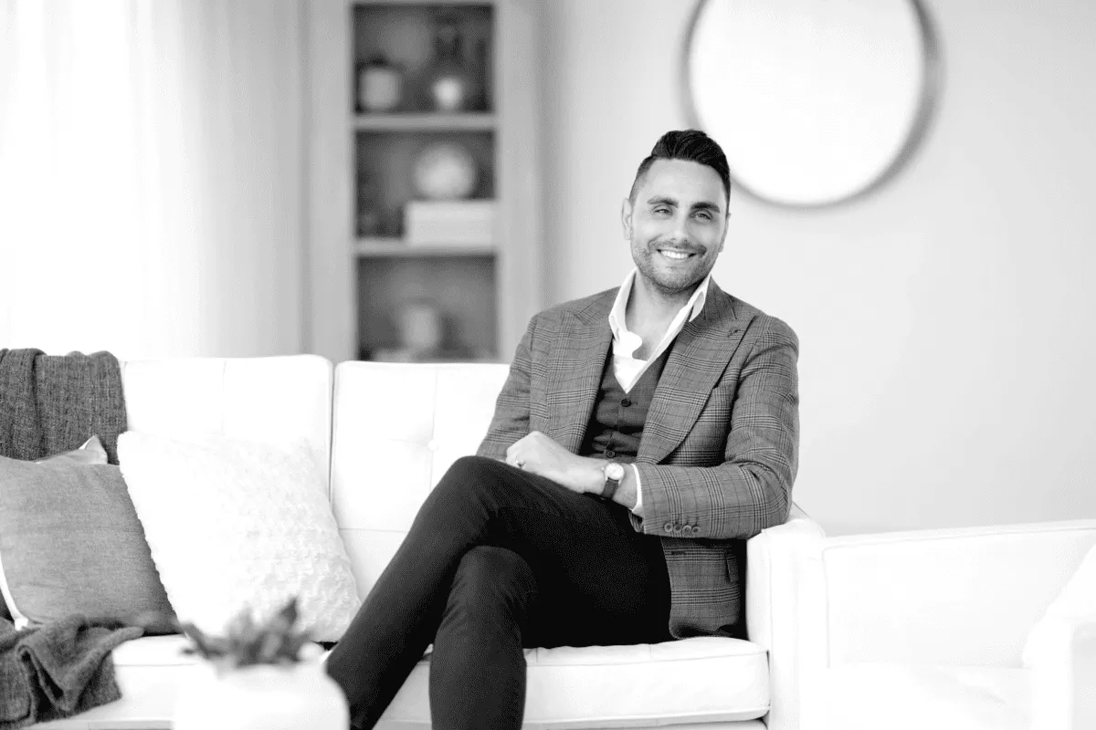 Smiling man in a checkered blazer and dark pants sitting cross-legged on a white couch in a modern living room.