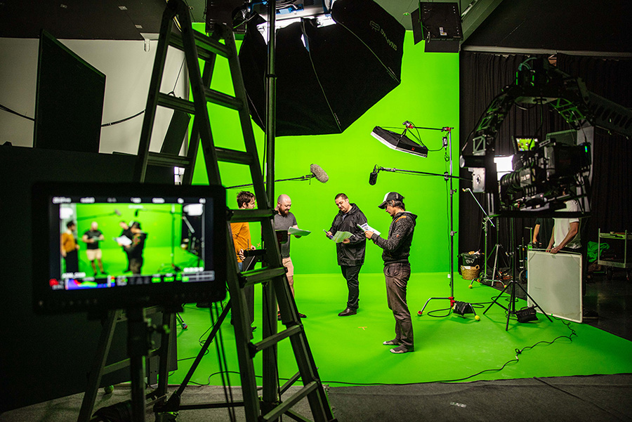 Four men reading scripts on a green screen studio set with cameras and lighting equipment.