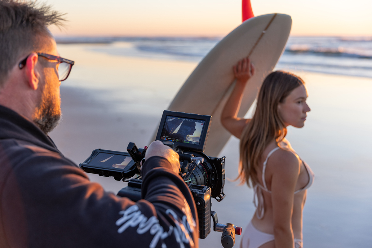 Man filming a woman in a bikini holding a surfboard on a beach at sunset.
