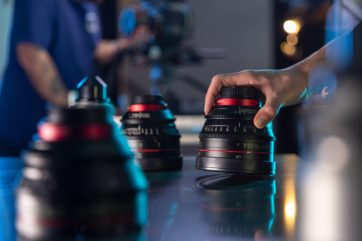 Hand adjusting a Canon camera lens on a reflective surface with other lenses and blurred background.