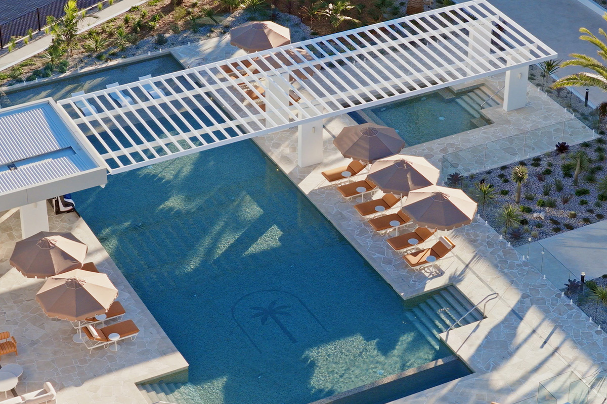 Aerial view of a blue pool with a palm tree design, lounge chairs, umbrellas, and white pergola.