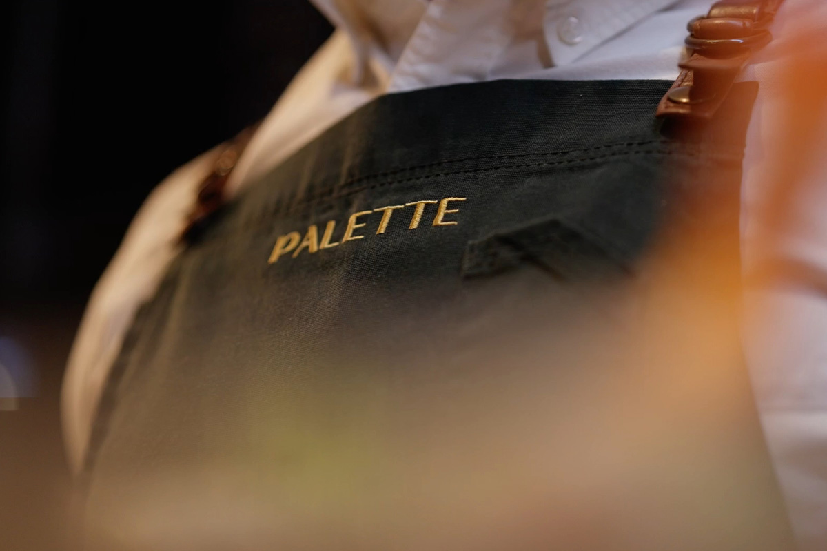 Close-up of a black apron with the word PALETTE embroidered in gold thread.