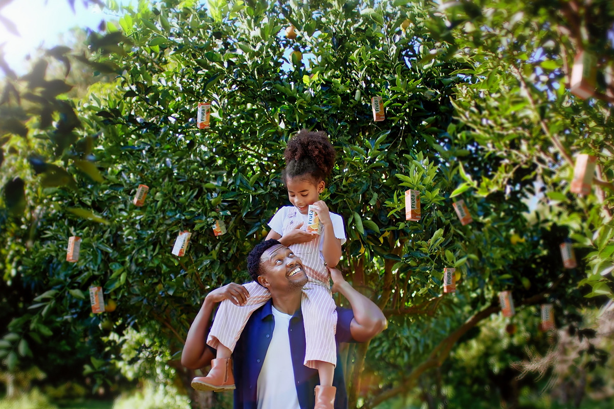 Father carries daughter on shoulders under a tree with hanging juice boxes in the branches.