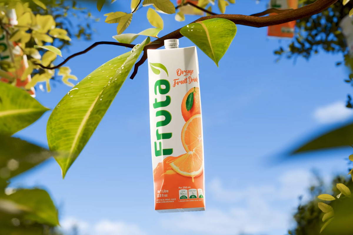 Carton of Fruta orange fruit drink hanging among green leaves under a clear blue sky.